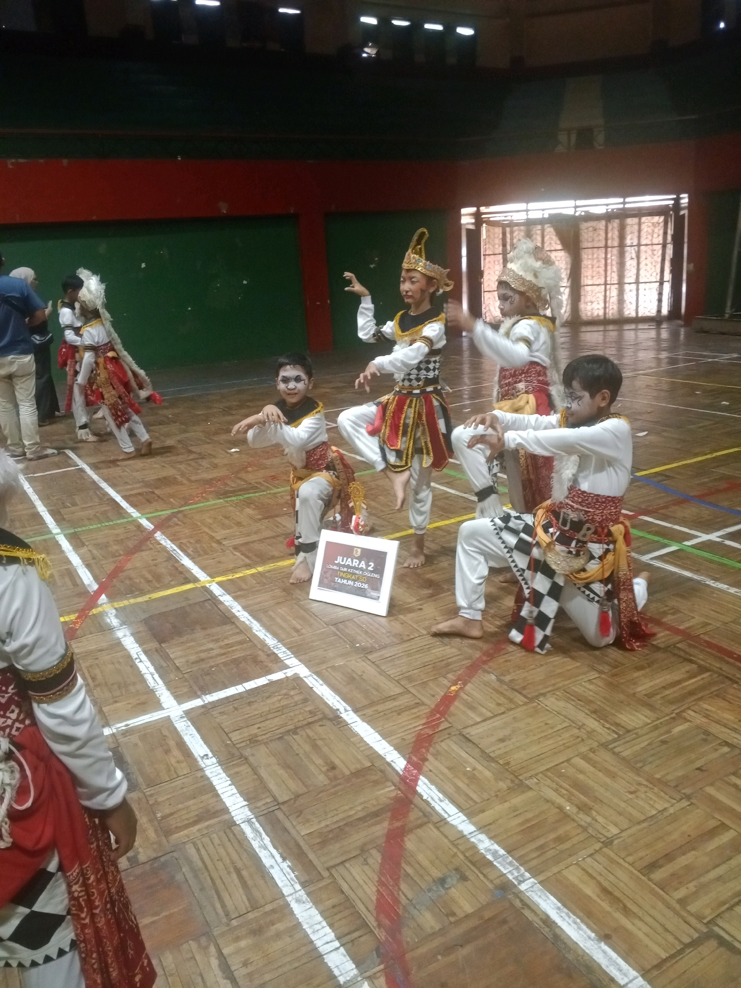 In the gymnasium of the SMP N 1 Wuryorejo, Indonesia, children in traditional Indonesian attire perform a dance. The participants, wearing white long-sleeved shirts, grey pants, and ornate headdresses, are adorned with sashes and patterned fabrics in red, gold, and black. Some have face paint, including dark eye makeup. The gymnasium floor is made of wooden planks marked with white and colored lines, indicating a court. In the foreground, a boy kneels with his hands positioned in front of him, while another boy in the background stands with his arms outstretched. A white sign with Indonesian text, reading "JUARA 2," is visible near the kneeling child. In the background, spectators are partially visible, with a green and red wall and tiered seating in the upper portion of the image. The overall scene captures a cultural performance taking place in a school setting.
