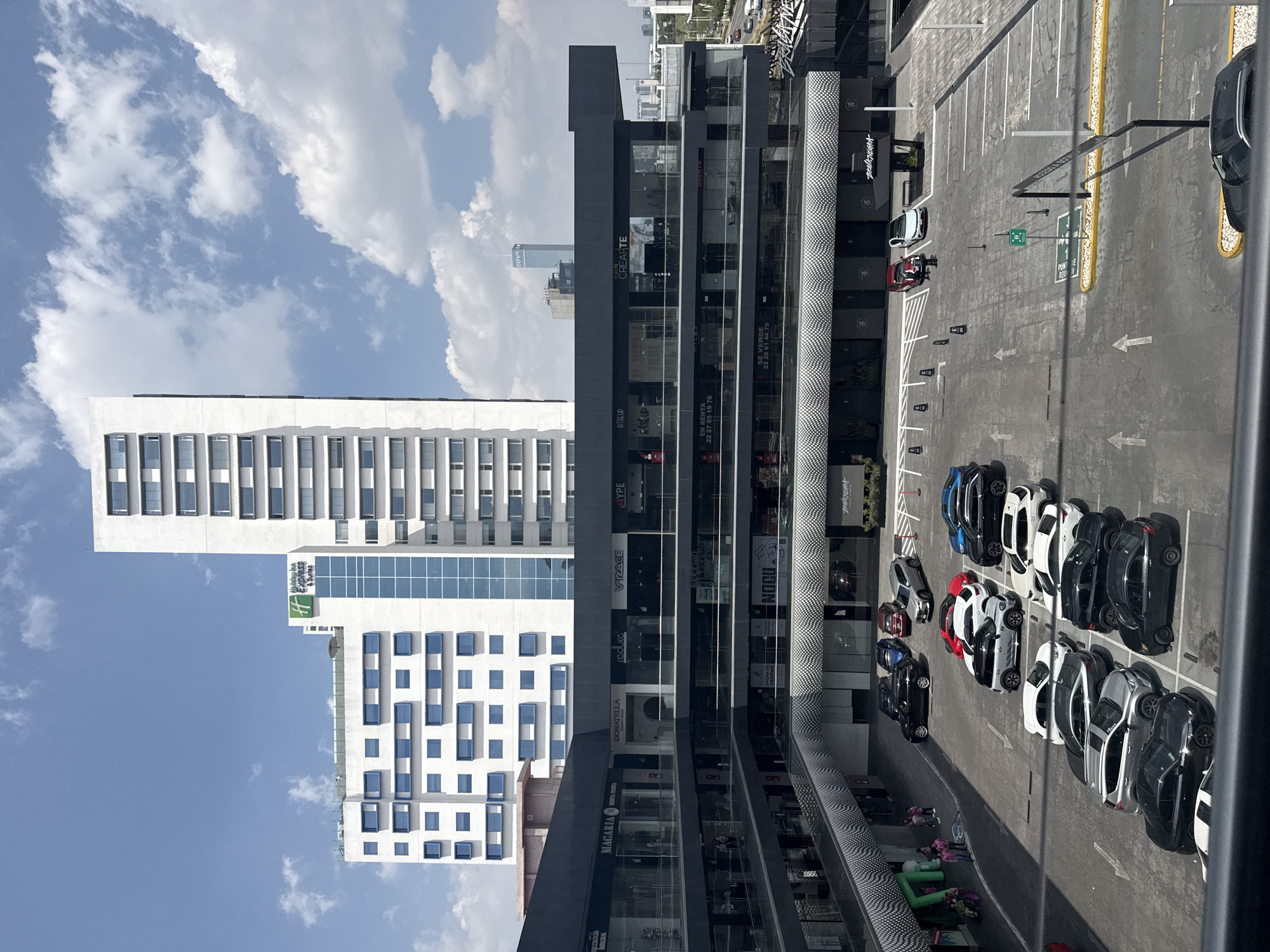 The image captures an urban scene in San Bernardino Tlaxcalancingo, Mexico. Several modern buildings dominate the view, including a Holiday Inn Express and a multi-story retail and office complex. The retail building showcases various businesses, with signage visible for companies like "MAGARIA," "DONATELLA," and "NOGU." Parking is adjacent to the structure, with a significant number of black, white, silver and blue vehicles parked in marked spaces. The sky features scattered clouds, indicative of fair weather.