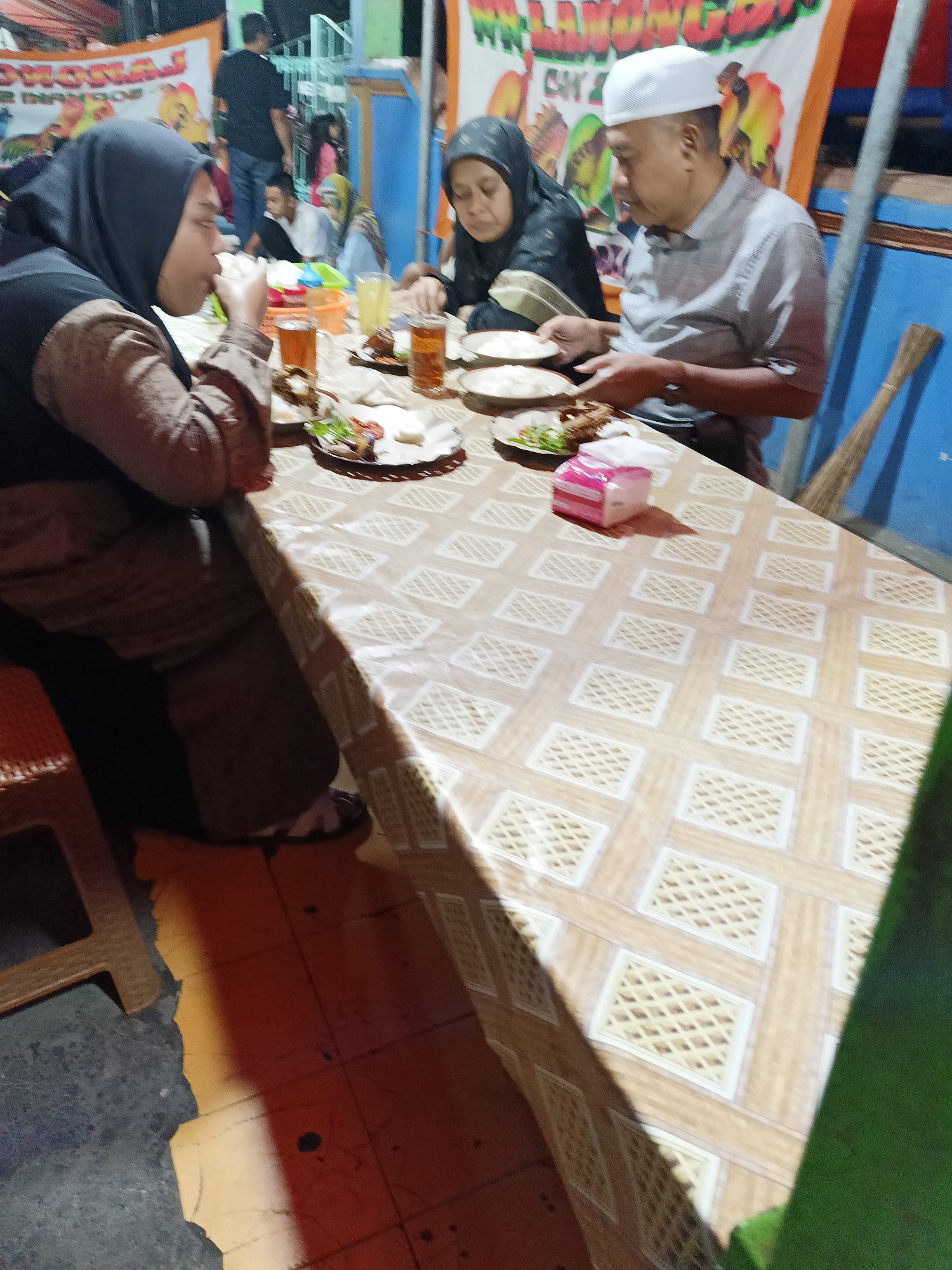 A group of people are seated at a table, eating a meal in what appears to be an outdoor setting. The table is covered with a patterned cloth, and there are plates of food, glasses of drinks, and a tissue box on it. Three people are clearly visible. On the left, a woman wearing a hijab is eating. In the center, another woman wearing a hijab is looking down at her plate. On the right, a man wearing a white cap and a grey shirt is also eating. In the background, more people are visible, and there are banners hanging on a wall. The lighting suggests it is either evening or overcast. The scene is consistent with a meal being shared at a modest eatery or gathering in Kebondalem Kidul, Indonesia.