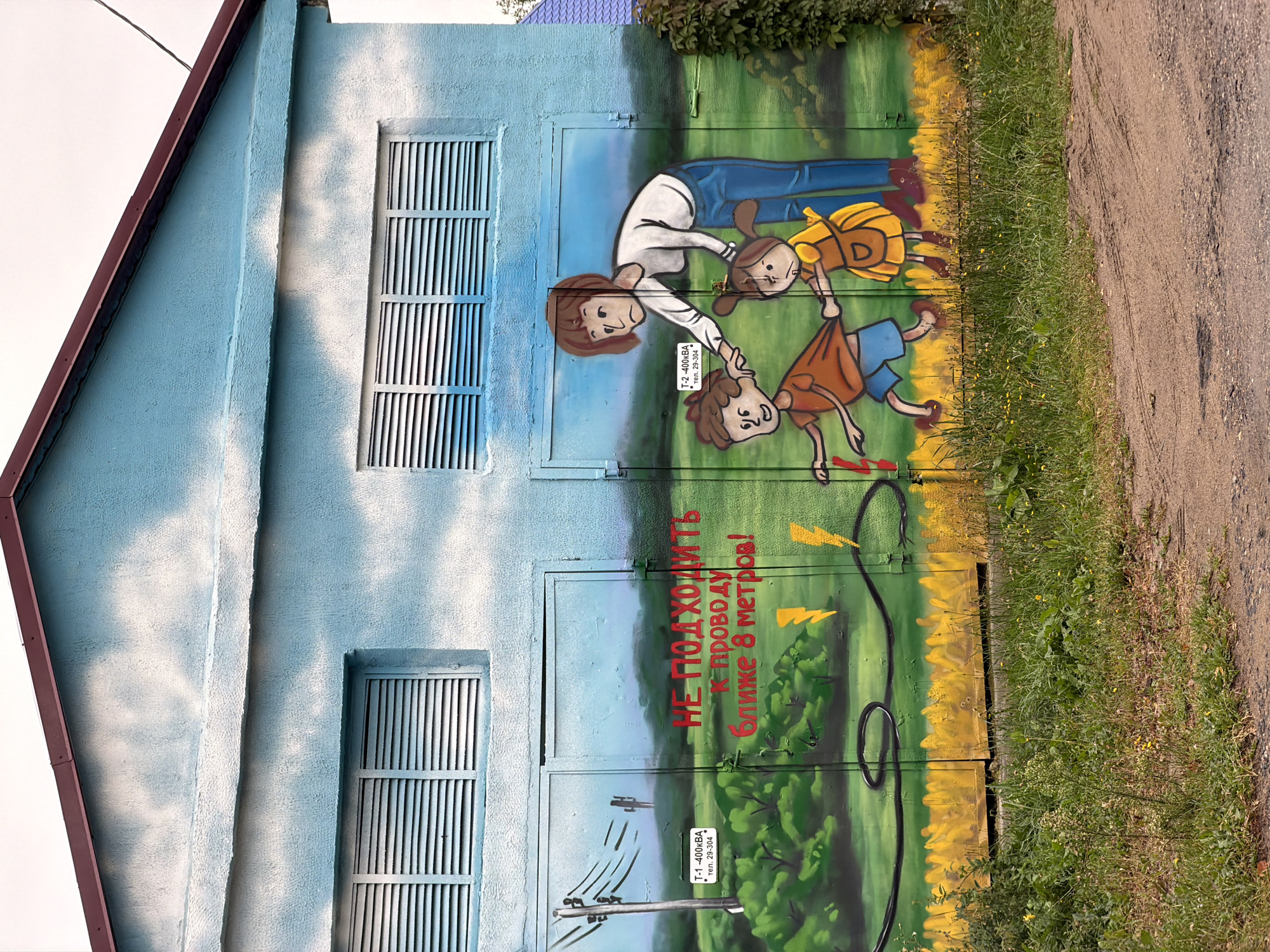 This image depicts a mural painted on the side of a light blue building, likely an electrical substation, in the city of Cherven, Belarus. The mural is a safety warning illustrated with cartoonish figures and text. On the left side of the mural, an electrical pole with wires is depicted, along with a sign that reads "T-1 -400 kVA tel. 29-304". The central part of the mural features red text that translates to "DO NOT APPROACH THE WIRE CLOSER THAN 8 METERS!". To the right of the text, a woman is shown holding back a boy and a girl, who appear to be playfully running towards a black electrical wire depicted on the ground, with yellow lightning bolts symbolizing danger. The background of the mural is a green landscape with a light blue sky. The building itself has two large shuttered windows above the mural. The area in front of the building is a dirt path with overgrown green grass and some yellow wildflowers.