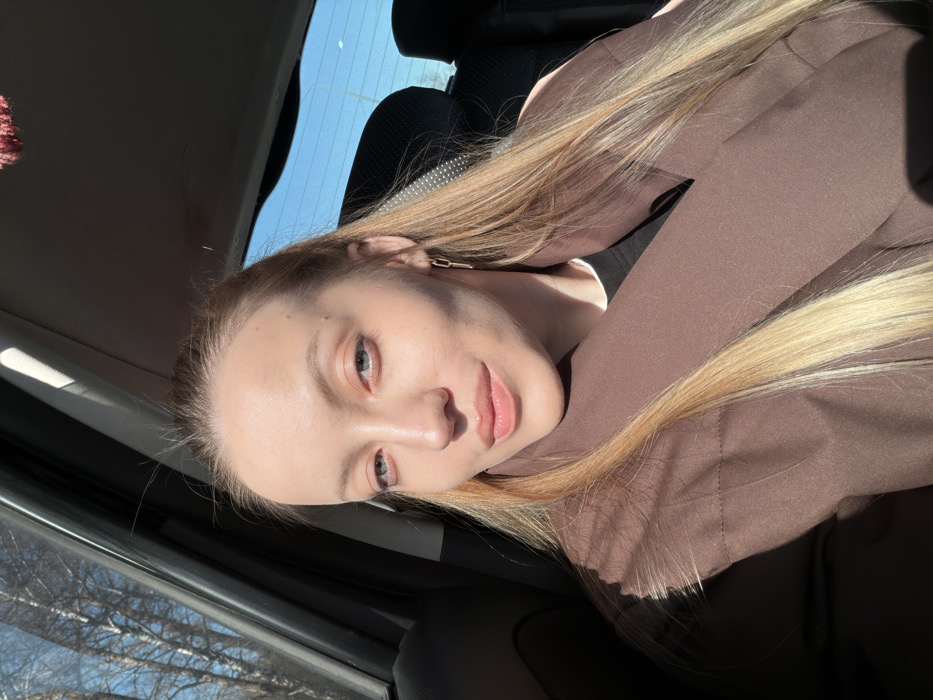This image depicts a single adult female seated within the passenger compartment of a vehicle, presumed to be a car, in Spas-Zaulok, Russia. The woman, with a fair complexion, long blonde hair, and light-colored eyes, faces forward with a slight smile and direct gaze. She is wearing a dark brown coat over a dark undergarment and a single gold-colored, elongated link earring is visible on her right ear. Strong natural sunlight enters the vehicle from the left, illuminating her and casting shadows across her face and jacket. Through the left side window, bare trees with thin trunks are visible against a bright blue sky. The vehicle's dark-colored interior elements, including a seat, a window frame, and a portion of the headliner, are discernible. A blurry, dark red decorative item is partially visible hanging from the interior roofline towards the rear. The rear window shows an unobstructed bright blue sky.