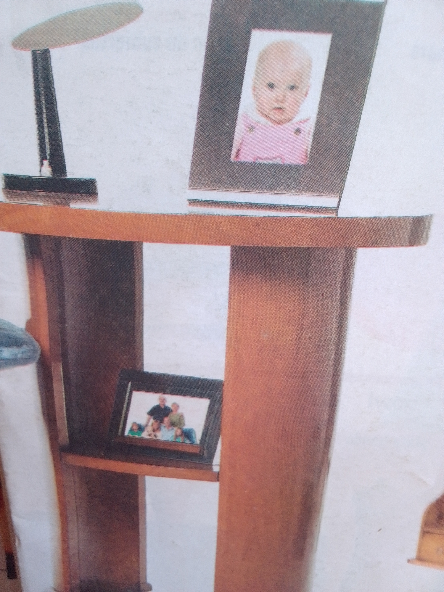 The image shows an indoor scene, likely a home or office, featuring a wooden console table with two framed photographs. On the top of the table, a framed photograph displays a close-up of a baby wearing a pink outfit. To the left of this photograph, a black desk lamp with a curved light fixture is positioned. On a lower shelf of the console table, a second framed photograph shows a family of six. The console table itself is made of dark wood and has a curved design. The background is a plain, light-colored wall. The scene does not provide any specific geographical markers to indicate the location within the city of La Tagua, Venezuela.