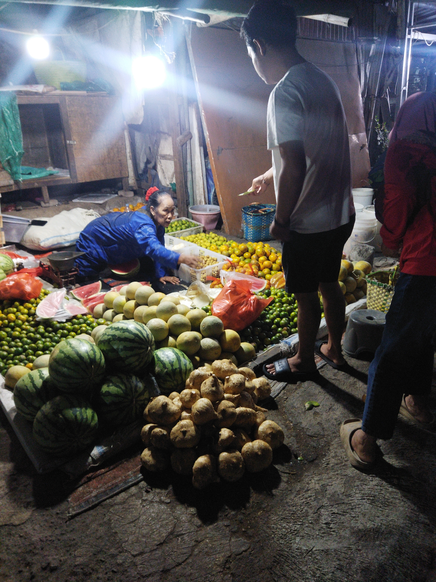 The image captures a nighttime scene at a market in Semarang, Indonesia, likely a street stall or an open-air market. The lighting is harsh and artificial, provided by bright overhead bulbs that create strong highlights and deep shadows.
In the foreground, a large pile of what appear to be yams or similar root vegetables sits on the ground. To the left of these vegetables, several watermelons are stacked. Beyond the watermelons, there are mounds of small, green citrus fruits, possibly limes or small oranges. Behind the citrus fruits, a woman wearing a blue garment and a red hair accessory is crouched down, attending to a basket of what seems to be more citrus fruits. She is interacting with a young man standing to her right, who is wearing a white t-shirt and dark shorts. He is holding a piece of green produce and appears to be in the process of purchasing or inspecting it.
Further to the right, a person wearing a red top and blue jeans stands partially in the frame. The ground is a rough, concrete or dirt surface, with some debris and water puddles visible, suggesting a humble market environment. Various containers, including plastic bags, baskets, and buckets, are scattered around the stall, holding produce. In the background, there are wooden structures and hanging tarps or cloths, typical of temporary market stalls. The overall impression is of a busy, albeit dimly lit, local market where fresh produce is being sold.