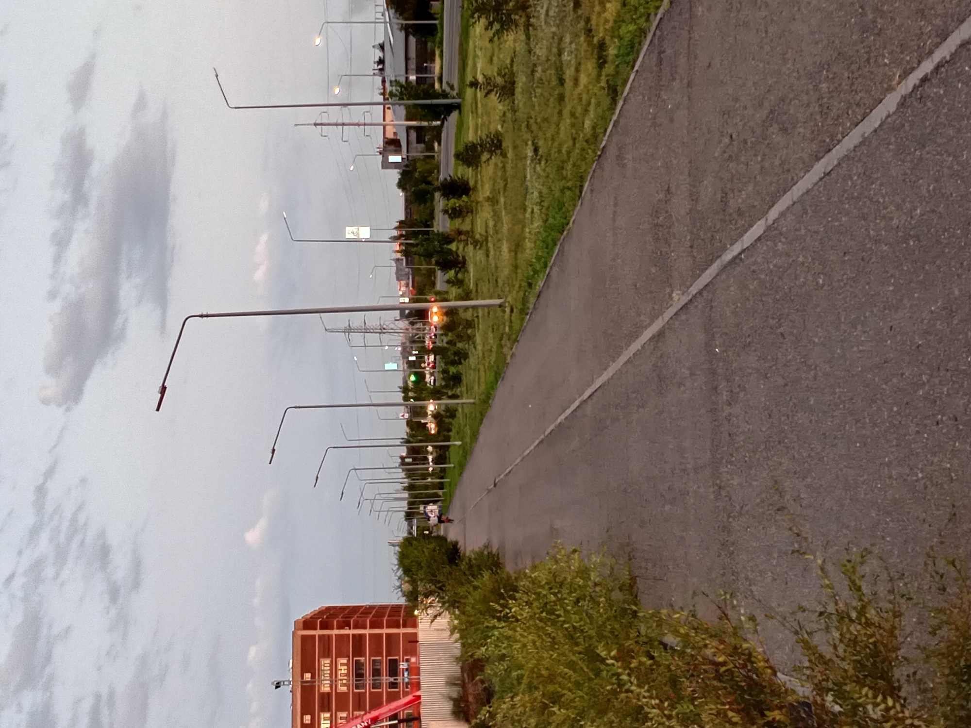 This image depicts a scene in the city of Semey, Kazakhstan, likely near a suburban or industrial area. A paved pathway, presumably for pedestrians and cyclists, runs diagonally across the frame from the lower left to the upper right. To the left of the pathway, dense green bushes and foliage line the edge. Further left, a tall, red brick building with multiple windows is visible, partially obscured by a construction fence and a red crane.
To the right of the pathway, a grassy area is interspersed with small trees and larger trees further in the background. A road runs parallel to the pathway, with vehicles visible, including cars with illuminated headlights. Numerous streetlights and utility poles are spaced along the right side of the road. The sky is overcast with scattered clouds, suggesting it may be late afternoon or early evening. A solitary cyclist is present on the pathway, moving away from the viewer. The overall impression is of an urban environment undergoing development or expansion, with a focus on infrastructure for transportation and public spaces.