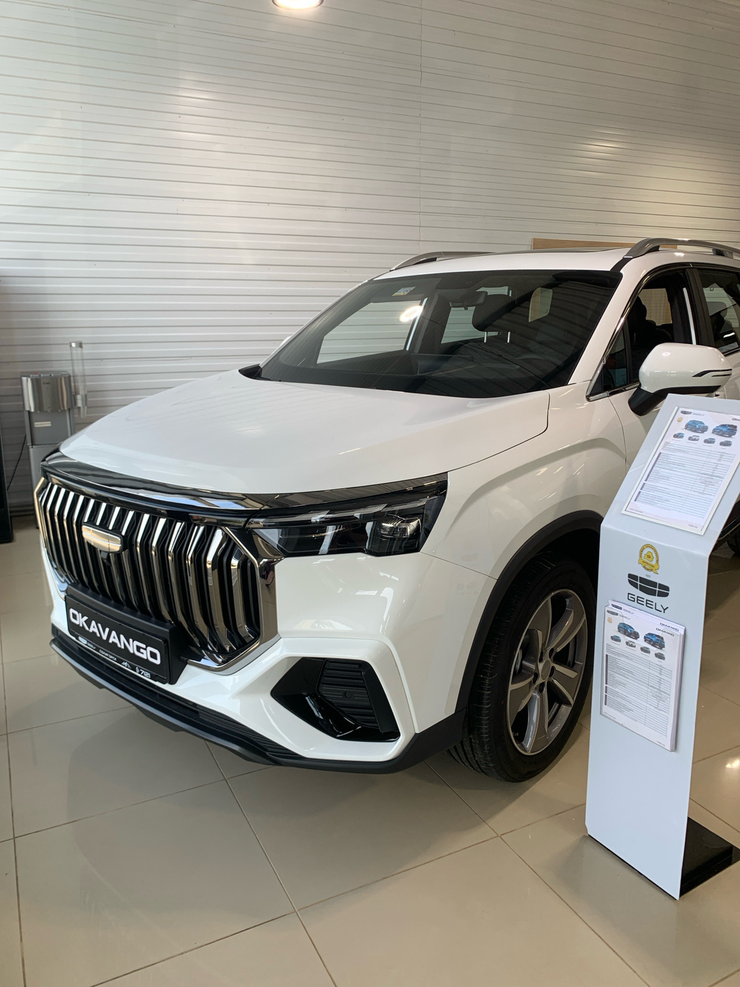 A white Geely Okavango SUV is parked indoors, likely in a car dealership, in Чыжэвіцкі сельскі Савет, Belarus. The car is angled towards the camera, with its front prominently displayed. The vehicle features a large, chrome grille with vertical slats and the "OKAVANGO" nameplate. The headlights are sleek and modern. A white display stand with information about the Geely brand and models is positioned to the right of the car. The background consists of a light-colored, corrugated wall and a tiled floor. No people are visible in the image.
