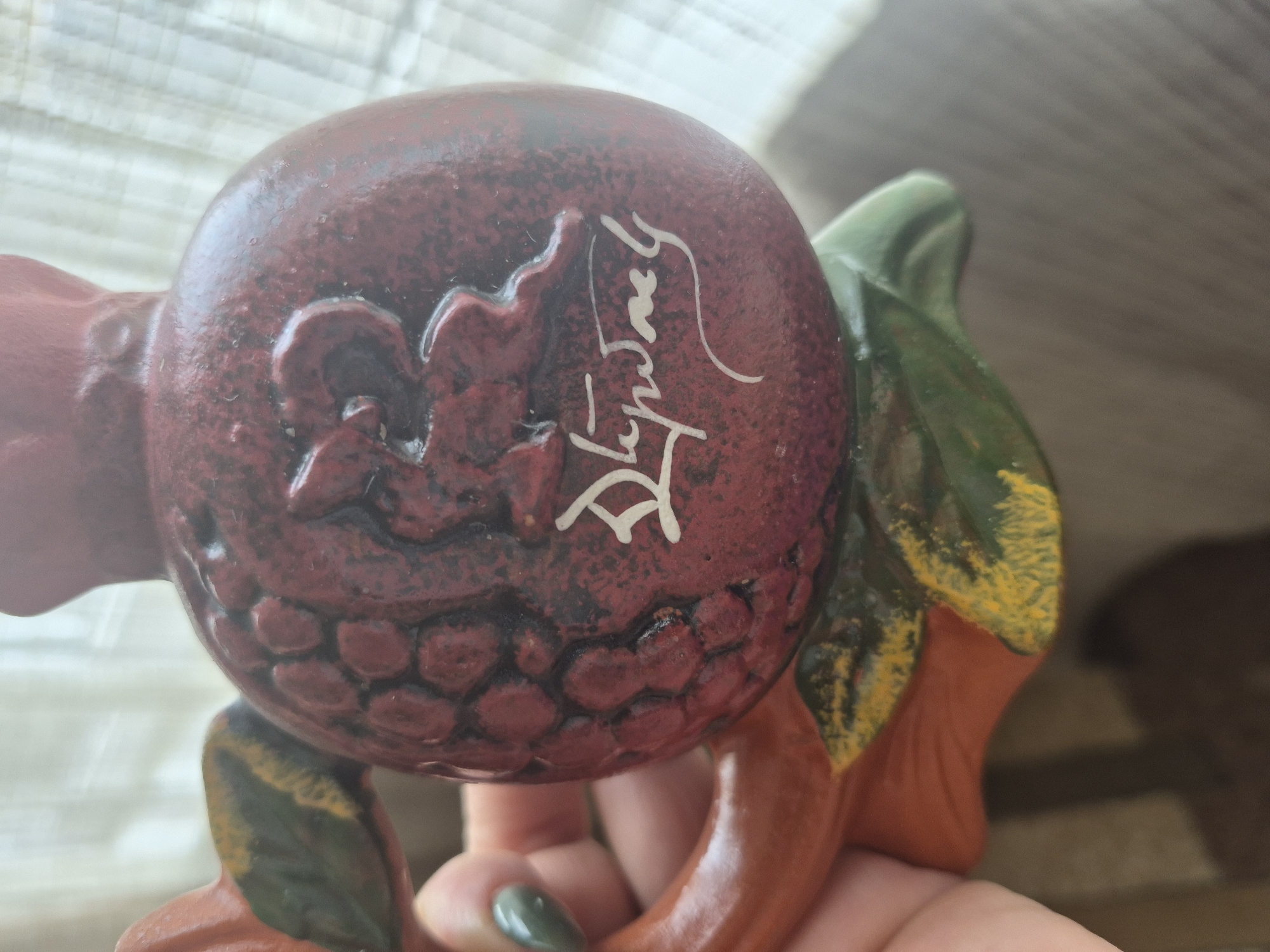 The image shows a close-up view of a ceramic pomegranate figurine, likely a decorative item. The figurine is primarily red with textured details mimicking the seeds of a pomegranate. There is a stylized depiction of a rabbit or hare in raised relief on one side of the pomegranate. Below the relief, there is white cursive script, possibly a signature or inscription. The figurine also features green and orange accents, possibly representing leaves or a stem. A human hand with painted fingernails is visible on the left side of the frame, holding the figurine. The background is out of focus, showing a soft, light-colored curtain or fabric. There is no discernible location in the city of Vanadzor, Armenia depicted in the image.