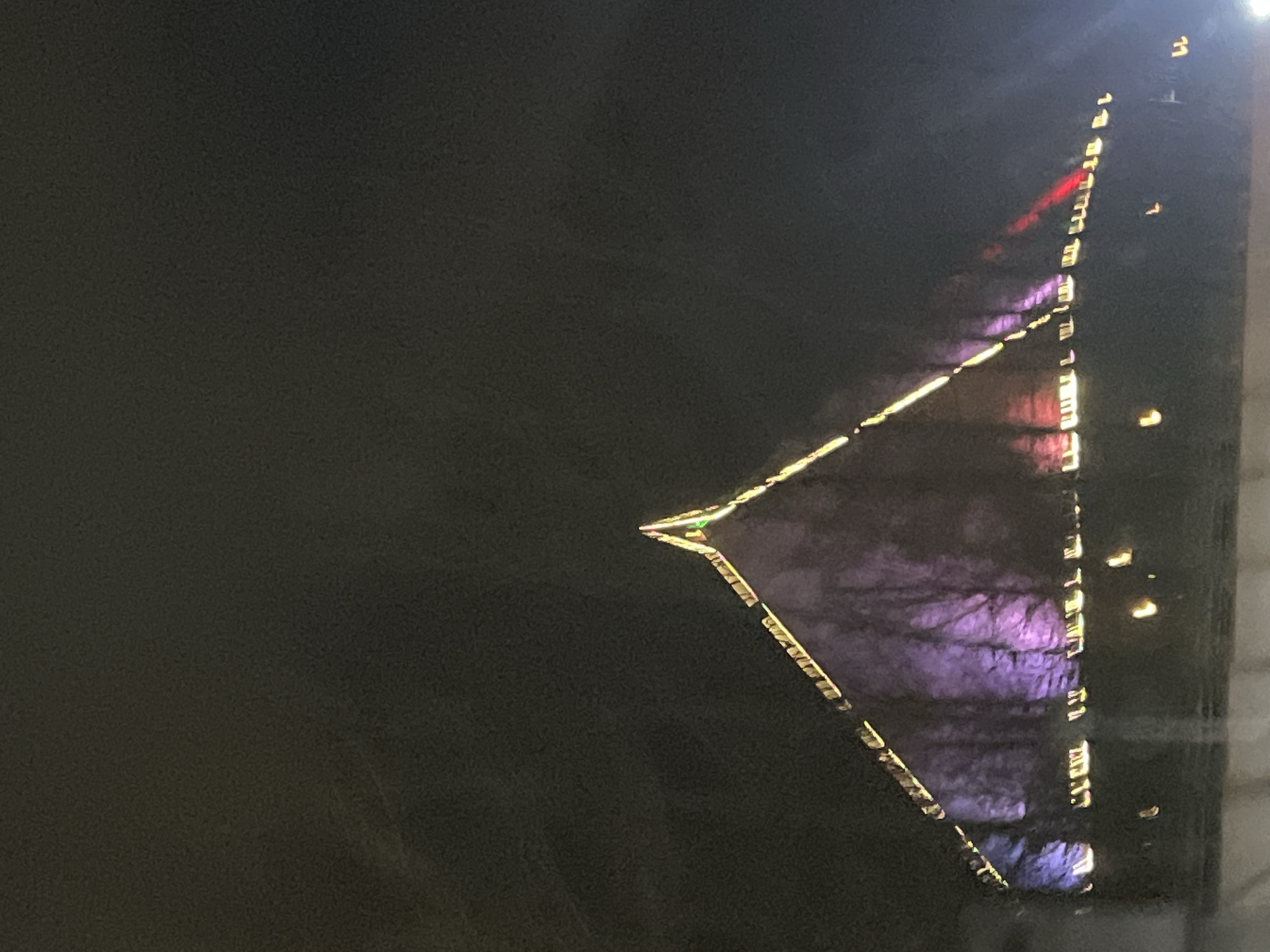 A night shot shows a large triangular building with illuminated edges in Verin Ptghni, Armenia. The building's reflection is visible, casting purple hues. There is a dark area in the left portion of the image.