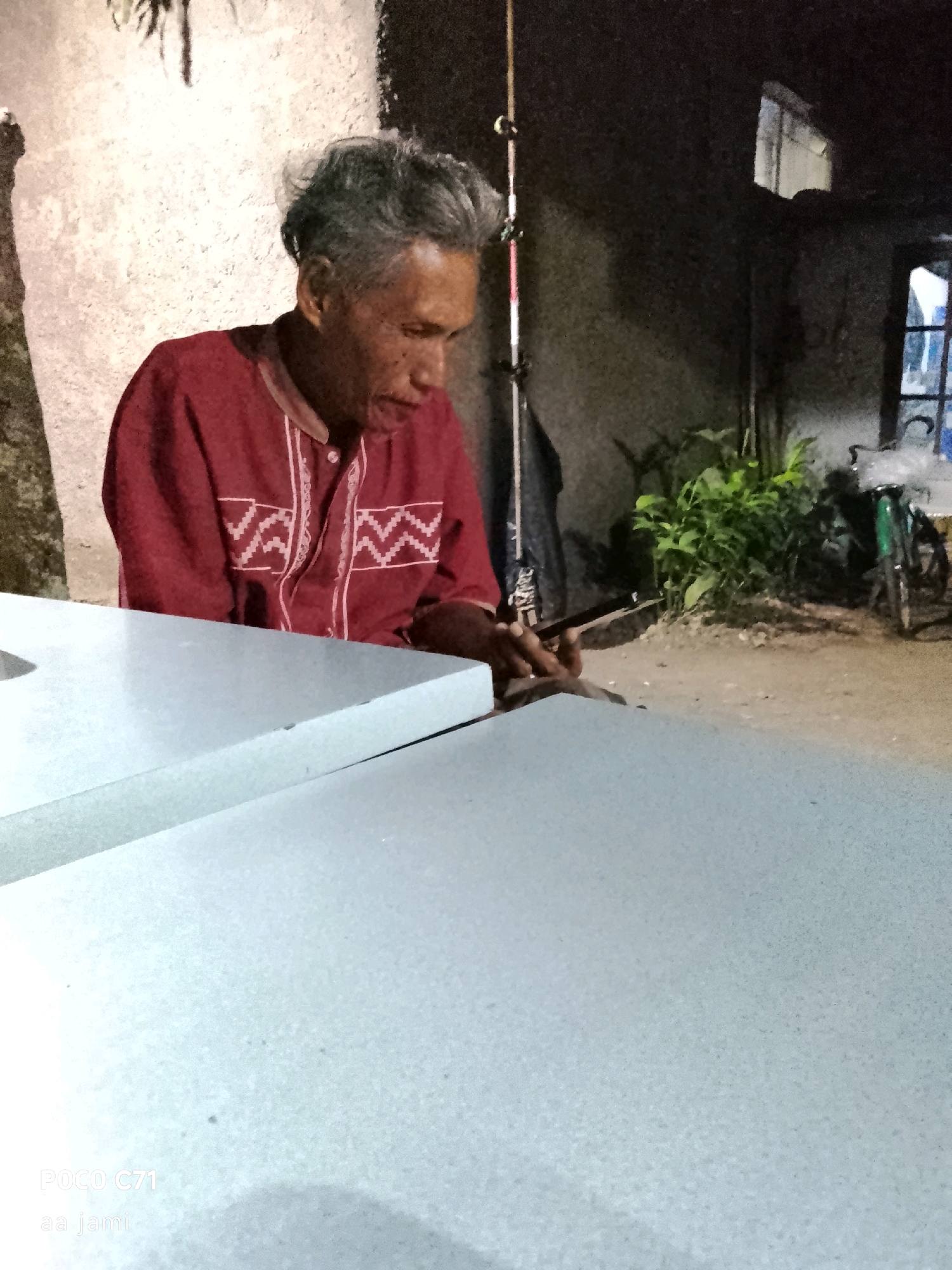 An elderly man with graying hair and wearing a red shirt with white trim is seated at a light-colored table. He is looking down, holding a small, dark object, possibly a phone or a tool, in his hands. The background suggests an outdoor or semi-outdoor setting at dusk or night, with a rough textured wall, some greenery, and a bicycle visible. The scene appears to be in Tasikmalaya, Indonesia, based on the timestamp "POCO C71 aa jami" which likely refers to a phone model and potentially a location tag or user name.
