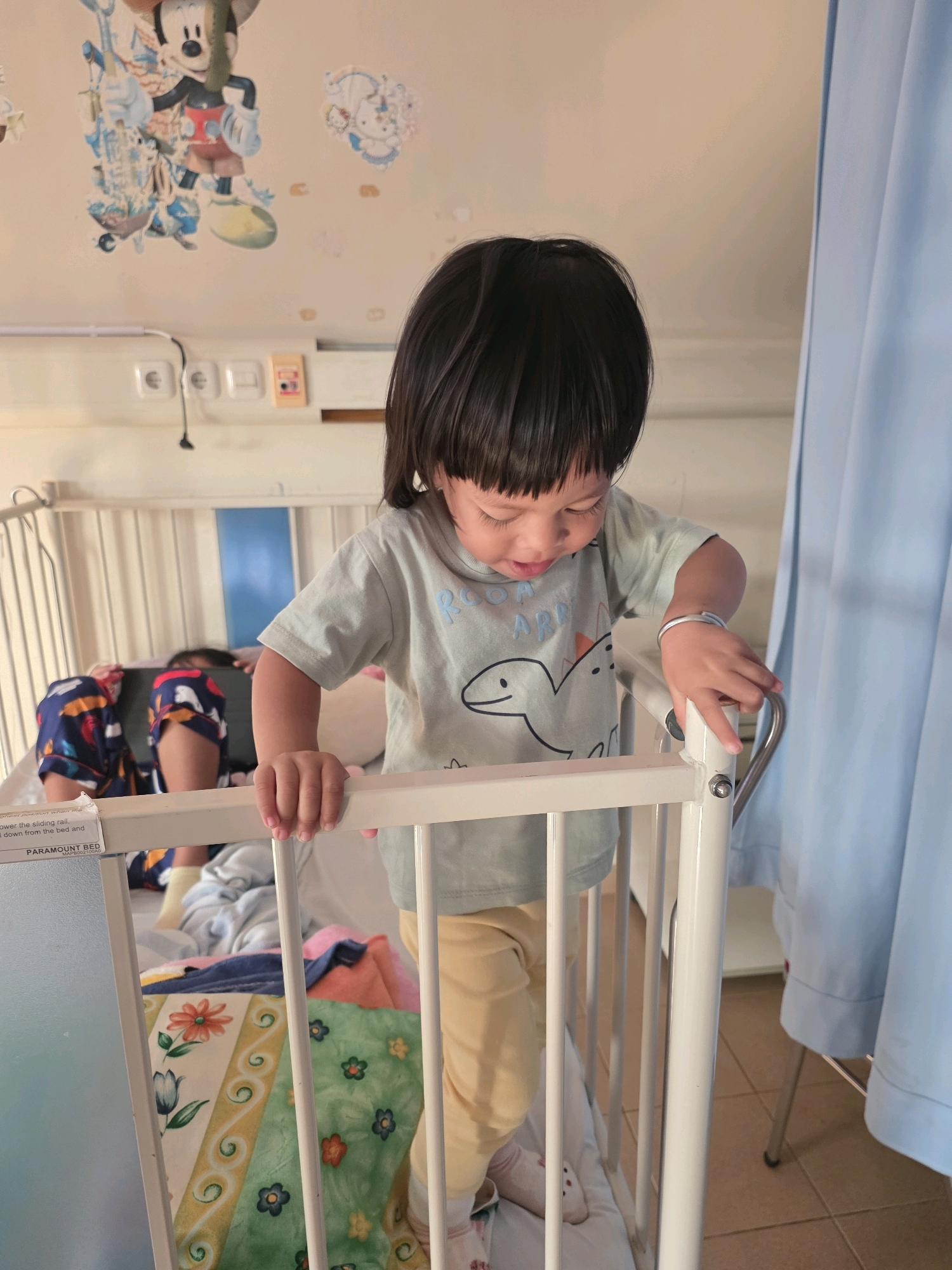 The image depicts a young child standing in a hospital crib, leaning over the rail and looking down. The child has dark hair and is wearing a light blue t-shirt with a dinosaur graphic and light yellow pants. Another child is visible lying down in the crib in the background, wearing pajamas with a colorful pattern. The crib has white metal railings and is adorned with a blanket featuring floral patterns. In the background, there are light blue curtains, electrical outlets on the wall, and cartoon decals of characters like Mickey Mouse. The floor appears to be tiled. The scene is likely in a hospital room in Tanjung Pinang, Indonesia, given the presence of hospital equipment and the general setting.