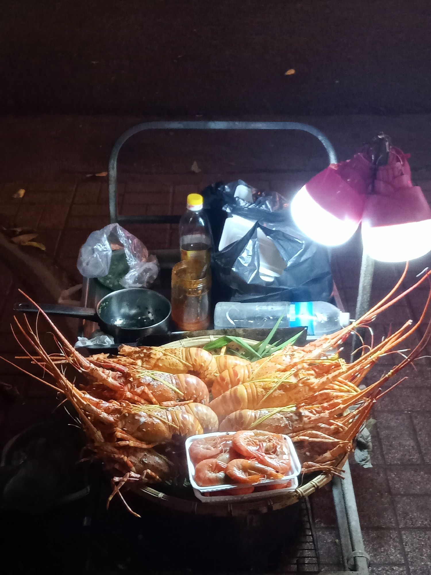 The image captures a vibrant street food stall scene at night, likely in Nha Trang Ward, Vietnam. The central focus is a large display of freshly cooked seafood, primarily several whole spiny lobsters (or similar large crustaceans) with long antennae, arranged in a woven basket or tray on a metal cart. The lobsters are reddish-orange, indicating they are cooked, and some appear to have steam rising from them.
Next to the lobsters, in a clear plastic container, is a portion of cooked pink shrimp. Surrounding the food display are various items essential for a street food vendor: a small metal pot or pan, a clear plastic bottle with a yellow cap containing what appears to be cooking oil or sauce, a clear plastic water bottle, a plastic bag containing green vegetables or herbs, and a black plastic trash bag.
The stall is illuminated by two bright, portable lights with pink/red casings, which cast a strong glow directly onto the seafood, highlighting its color and texture against the dark background. The overall setting suggests an active night market or street food environment. No people are visible in the frame, and there is no discernible text on any of the objects. The metal framework of the cart or stand is visible, hinting at the portable nature of the vendor's setup.