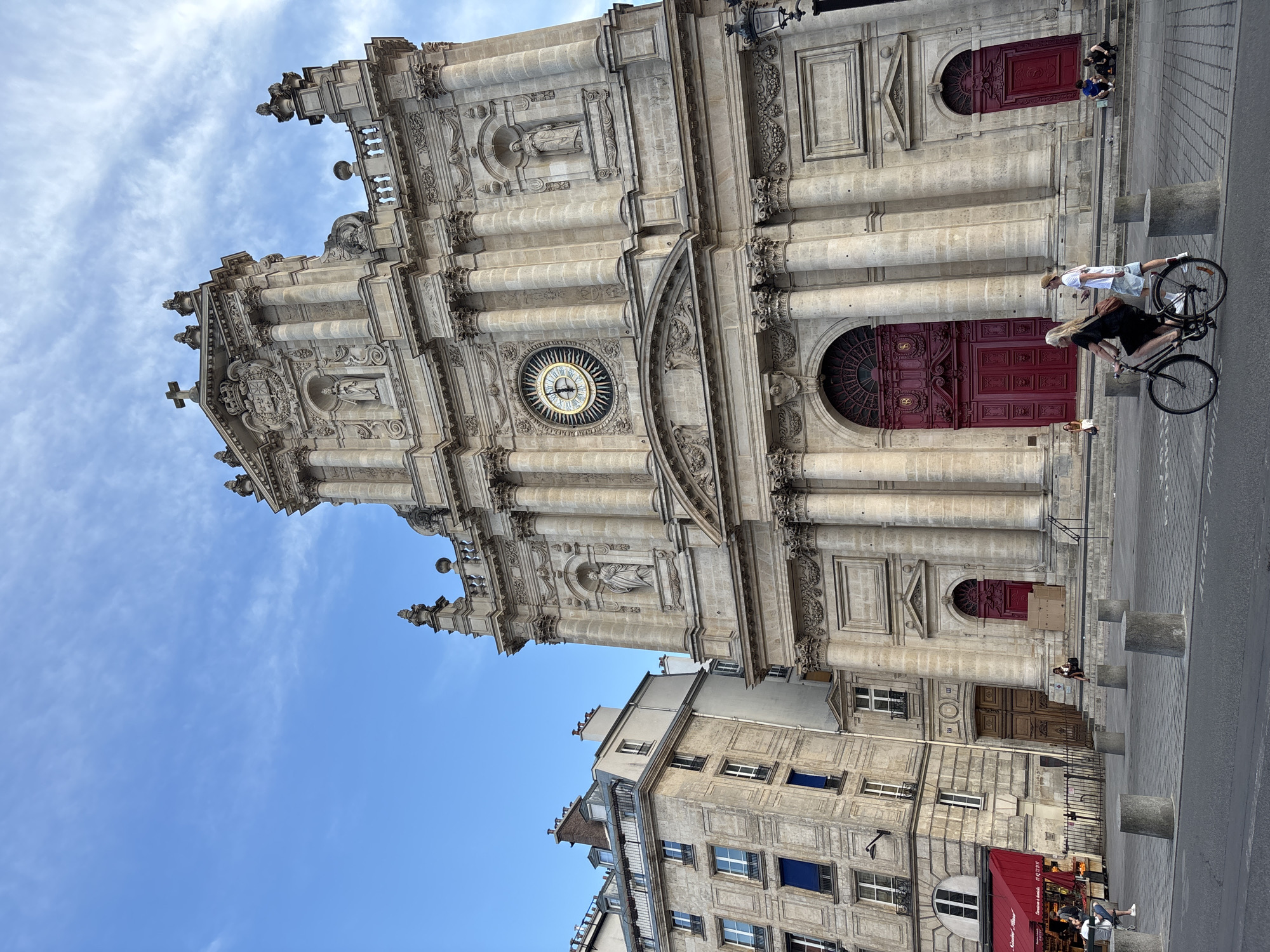 The image depicts the exterior of the Église Saint-Roch, a Roman Catholic church located in the 1st arrondissement of Paris, France. The church's facade, constructed of ornate stone, features classical architectural elements such as columns, statues in niches, and a large circular clock above the main entrance. The clock face has Roman numerals and ornate hands indicating the time is approximately 11:55.
To the left of the church, a multi-story building with traditional Parisian architecture stands, featuring multiple windows and a red awning at street level, suggesting a commercial establishment.
In the foreground, a paved plaza area is visible with several cylindrical concrete bollards. A woman is riding a bicycle, wearing a black dress and carrying a dark shoulder bag. She is moving from right to left across the frame. To her right, a man wearing a light-colored t-shirt and shorts walks in the same direction.
Several other individuals are present. A woman sits on the steps leading up to the church entrance on the right side. A group of people, appearing to be young males, are seated on the steps on the far right. Further back, near the entrance of the adjacent building, a woman sits on the steps, and another person is partially visible to the left of the red-doored entrance.
The sky above is blue with scattered white clouds. The lighting suggests it is daytime, possibly late afternoon given the direction of some shadows.