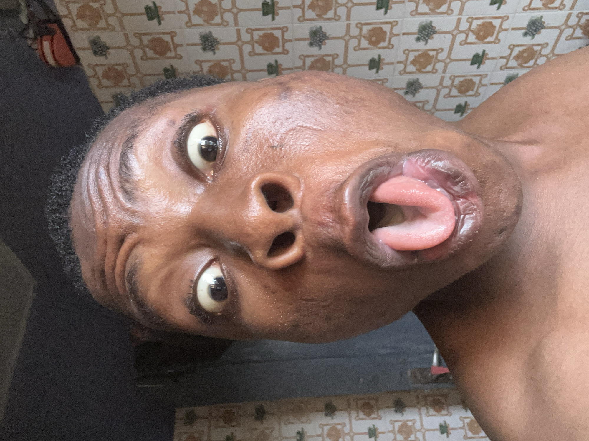 A close-up, angled view of a dark-skinned person's face with their tongue sticking out. The person has wide eyes and is looking directly at the camera. The background is a tiled wall. The location is not specified and cannot be determined from the image.