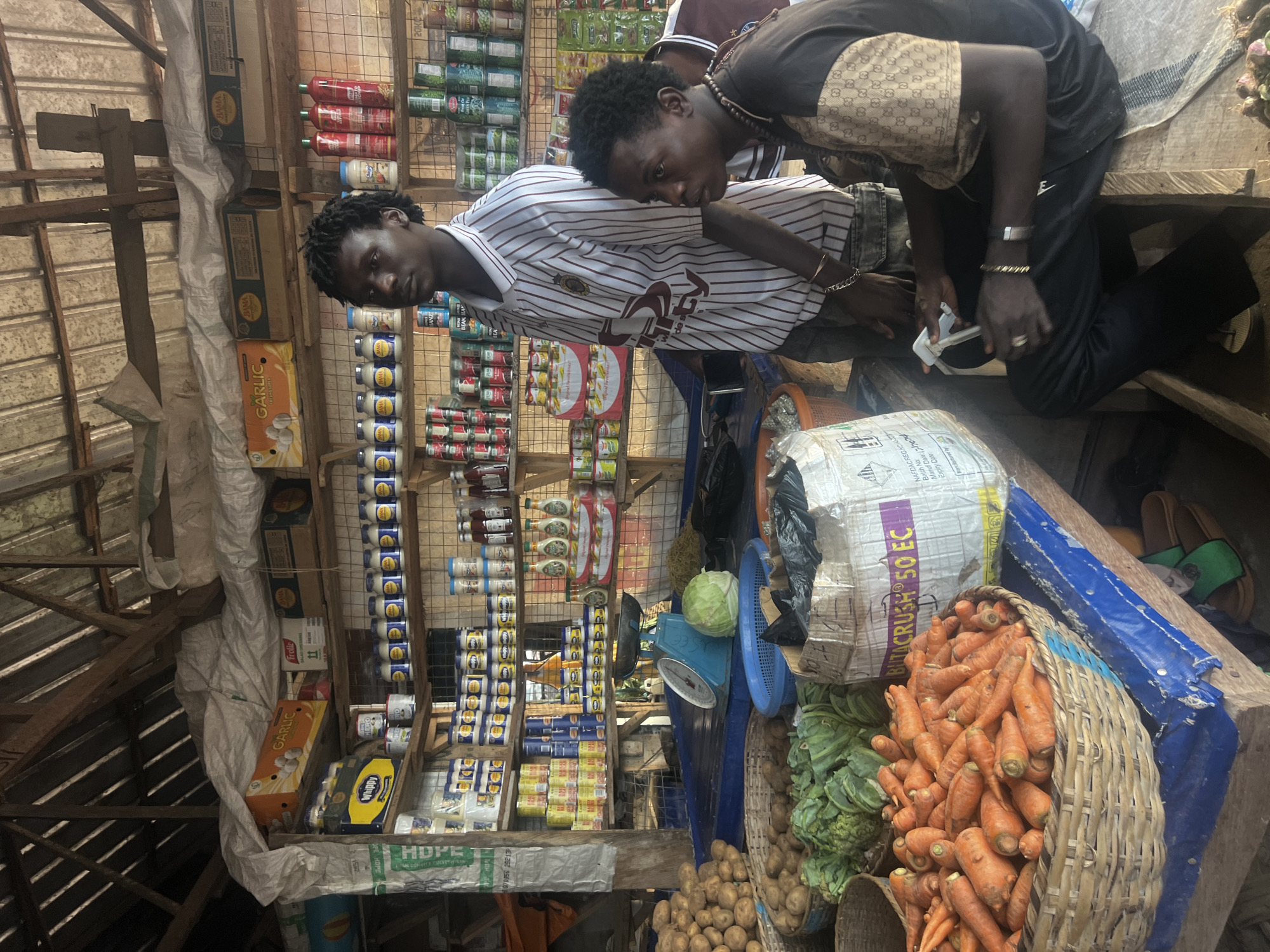 This is a street market scene in Oko Amakom, Nigeria. Two young men are present. The man on the right is seated on a wooden bench, wearing a black t-shirt with a pattern on the sleeves and black pants. He is holding a white object, possibly a phone or wallet, and is looking to his right. He is also wearing a necklace and bracelets. The man on the left is standing behind the seated man, wearing a striped t-shirt and dark pants. He is looking towards the camera.
The market stall is filled with various goods. In the foreground, there is a large basket of carrots and a basket of potatoes. There are also baskets of leafy green vegetables, a cabbage, and a scale. Shelves behind the men are stocked with canned goods, bottles of sauces and condiments, and packaged food items. The stall has a rustic wooden structure with a corrugated metal roof, suggesting an open-air or semi-enclosed market setting. A blue tarp is spread over some of the surfaces. Various bags and boxes are also visible, including a large white bag with text that appears to be in English. The overall impression is one of a busy, local marketplace.