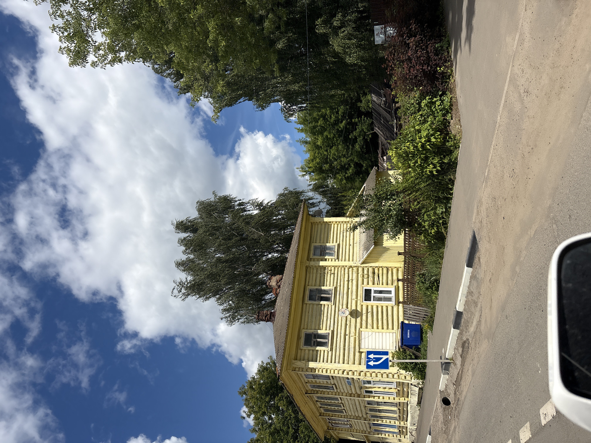 This is a vertical, eye-level, outdoor shot of a yellow, wooden, two-story building situated next to an asphalt road. The building is constructed from logs and has numerous windows with white frames. A blue sign with white arrows indicating a mandatory direction of travel (left and right) is visible in front of the building. A blue trash receptacle is also present near the building. The scene is framed by lush green trees and a bright blue sky with scattered white clouds. The lighting suggests it is daytime, likely sunny. The image has a rustic feel, possibly indicating a rural or historical setting. A black and white striped curb runs along the edge of the road. The scene appears to be in Pereslavl-Zalessky, Russia. No people are visible in the image.