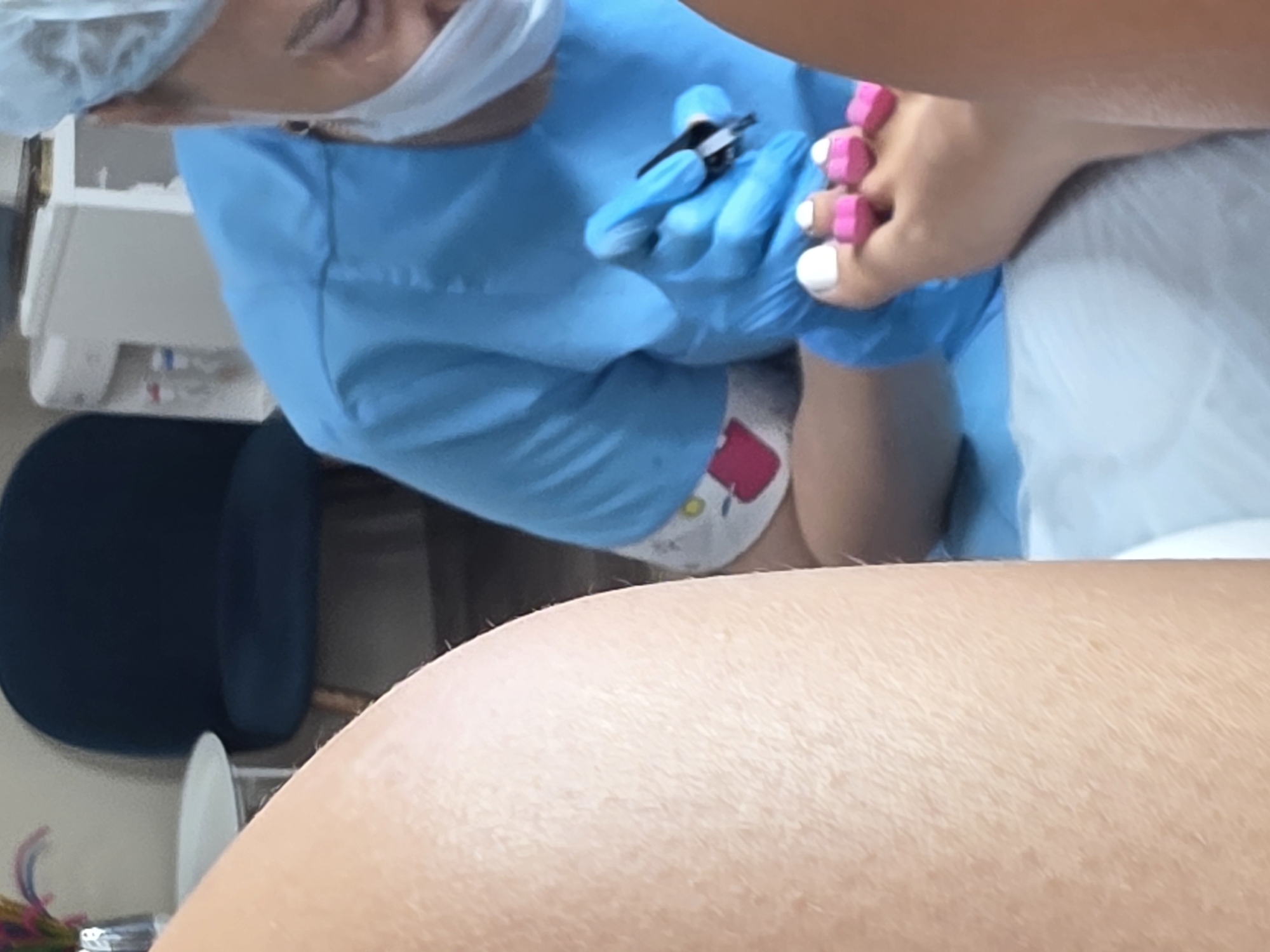 The image captures a close-up view of a pedicure service taking place, likely in a salon or spa environment. The primary focus is on a person's foot, with a technician, wearing blue gloves and a light blue medical scrub, working on the toenails. The toenails are painted white, and pink toe separators are in place. The technician is holding a tool, possibly nail clippers or a file, and is actively engaged in the process.
The background is slightly out of focus but reveals elements of the setting. A dark blue upholstered chair is visible, suggesting a seating area. To the left, a small white table with a vase containing colorful decorative items is present. Further in the background, a white appliance, possibly a water cooler or a piece of medical equipment, can be seen, along with a monitor.
The lighting appears to be artificial and bright, illuminating the area where the pedicure is being performed. The overall scene conveys a sense of personal care and beauty treatment.
The provided information states this image is located in Sochi, Russia. This suggests the salon or spa is situated within this Russian city.