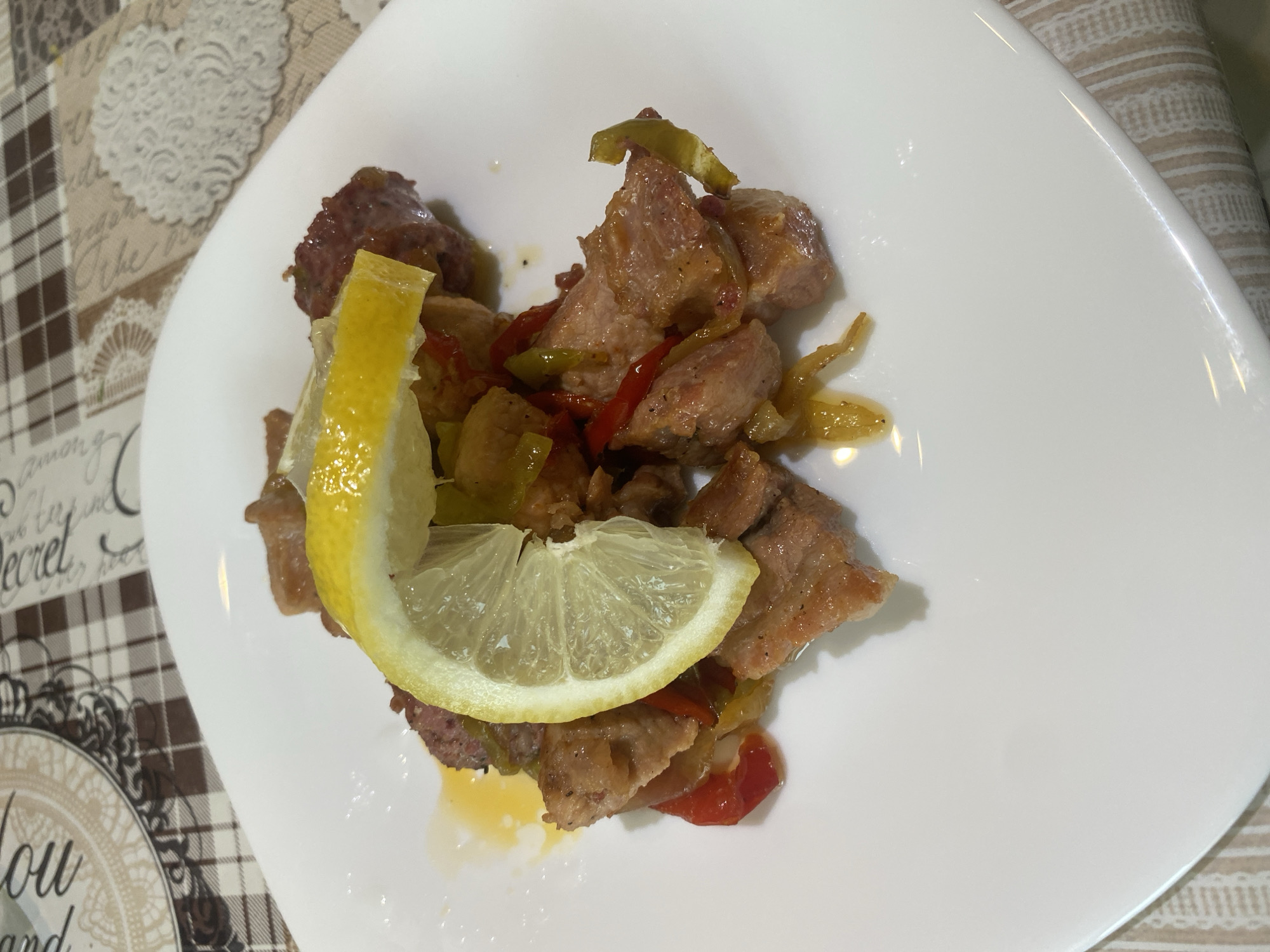 A close-up overhead shot displays a portion of cooked meat with vegetables and lemon on a white, elongated plate, set on a patterned tablecloth. The main dish consists of cubed pieces of light brown, cooked meat, possibly pork or chicken, interspersed with vibrant red and green bell peppers or similar vegetables. A thick wedge of fresh lemon is placed upright on the food, and a thin lemon slice rests horizontally below it, partially submerged in some cooking juices. The food appears to be glazed or sauced, contributing to its glossy appearance.
The white plate has a slightly irregular, sleek shape and reflects ambient light, indicating a smooth, shiny surface. It is positioned on a checkered tablecloth with a brown and beige pattern. Parts of the tablecloth feature ornate, lace-like designs and decorative script. Visible text on the tablecloth includes words such as "The secret", "among", "you", and "and". The lighting suggests an indoor setting. No people are visible. The image captures a meal ready to be consumed, offering a glimpse into a dining experience in Sliven, Bulgaria.