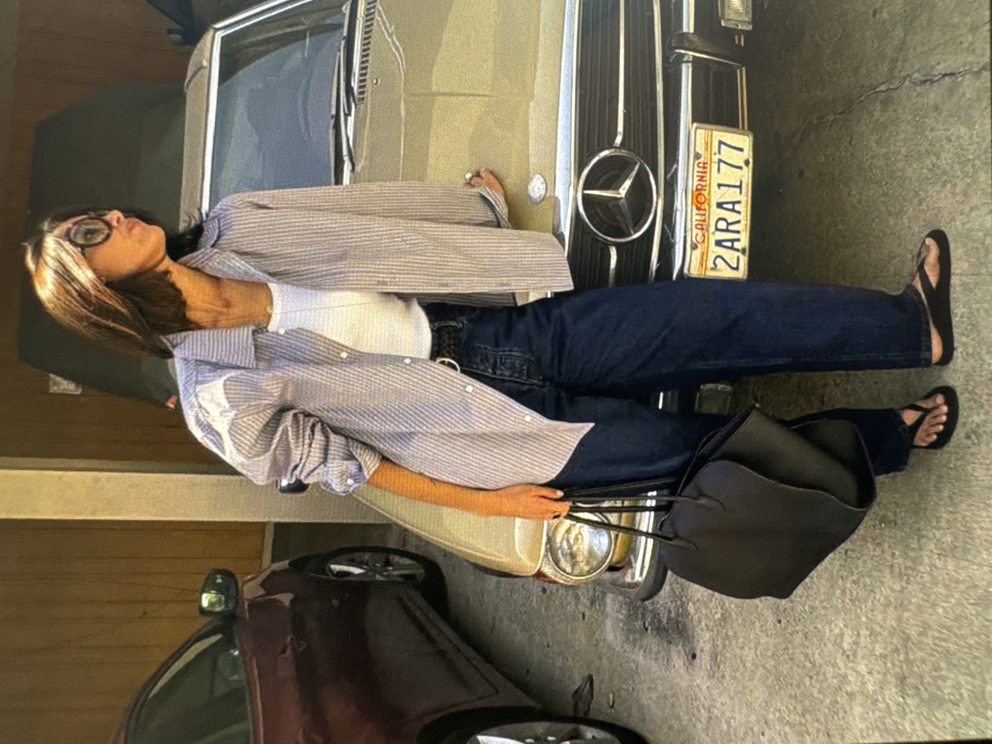 A low-angle shot shows a woman lying on the hood of a vintage yellow Mercedes-Benz. She is wearing a white tank top, a blue and white striped button-down shirt, dark wash jeans, and black flip-flops. She has on large, round sunglasses and her hair is a mix of dark brown and blonde. Her head is tilted back, and her eyes are closed, suggesting a moment of relaxation or repose. One hand rests on the hood of the car, near a chrome emblem. A large, dark leather tote bag sits beside her on the ground.
The car's front grille and a California license plate, reading "2ARA177", are visible. The background suggests an outdoor setting, possibly a driveway or garage, with another dark-colored car visible in the lower left corner. The lighting indicates it might be daytime, with dappled shadows suggesting sunlight. The overall mood is relaxed and casual.