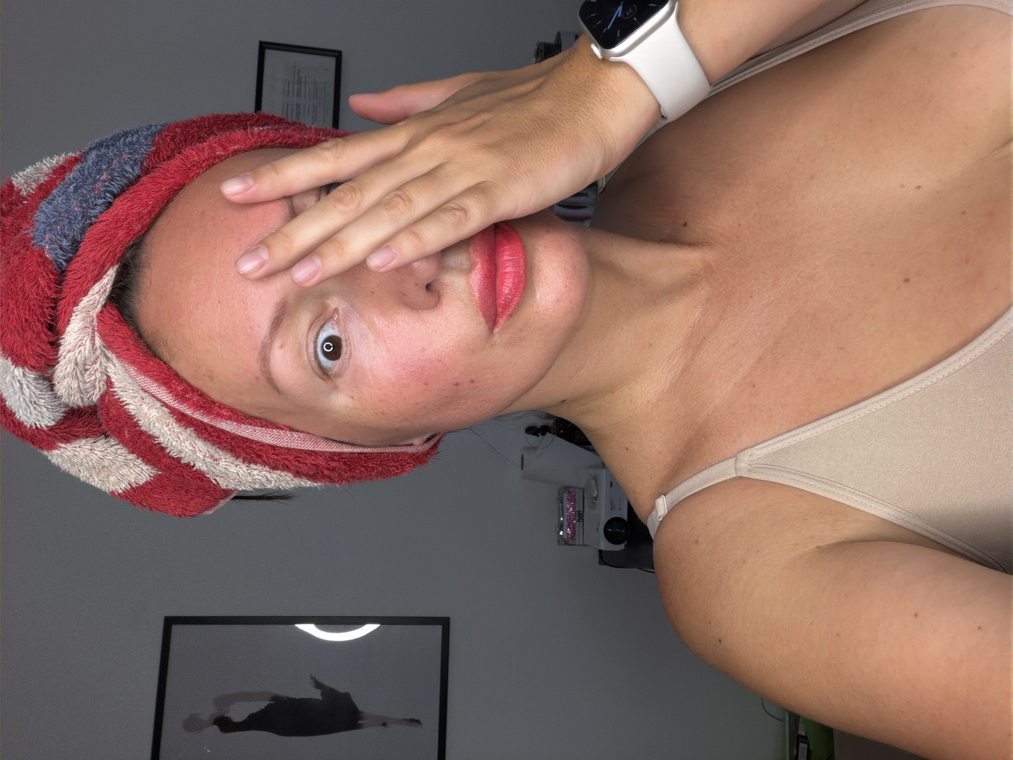 A woman with a red and white striped towel wrapped around her head and wearing a beige bra is touching her face with her right hand, obscuring part of her forehead and eye. She is looking directly at the camera with her left eye open. Her lips are painted red. She is wearing a white smartwatch on her left wrist. The background includes a framed black and white image of a silhouette of a woman in a dress on the left wall and a small white projector on a table in the background. The lighting appears to be artificial, likely from a ring light. The scene is indoors, and the location is not specified within the image or its context.