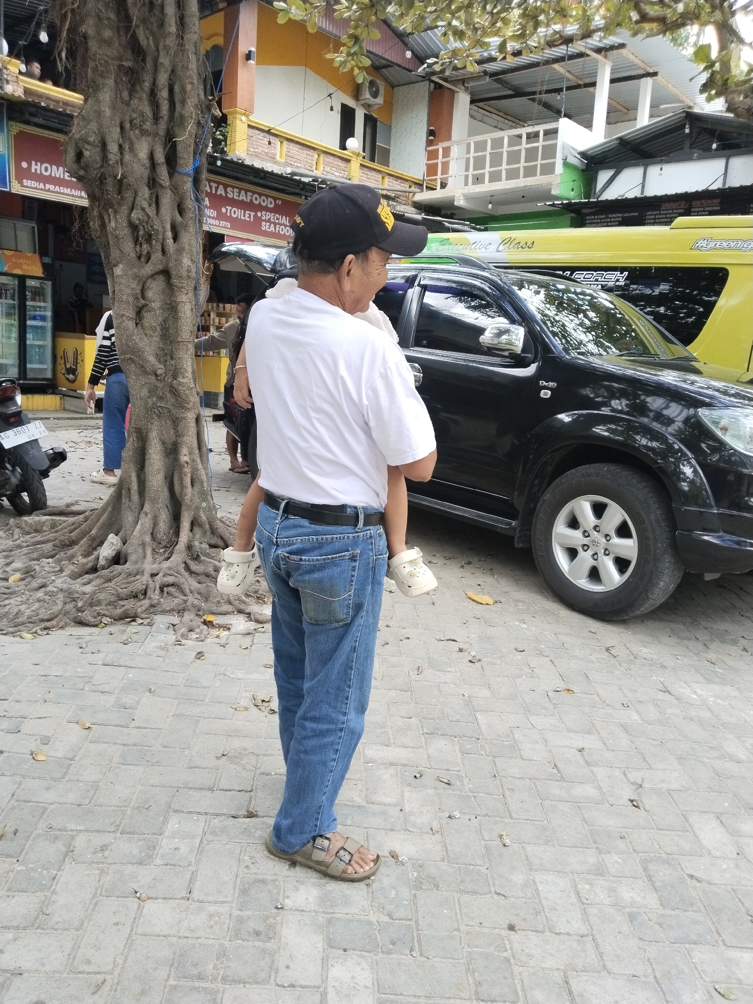 A man is carrying a small child in his arms in front of a building and a black SUV. The man is wearing a white t-shirt, blue jeans, and sandals. The child is wearing white shoes. The building appears to be a restaurant or shop, with signs in Indonesian. A large tree with exposed roots is on the left side of the image. The ground is paved with interlocking bricks. The scene appears to be an outdoor area on a street in Prigi, Indonesia.
