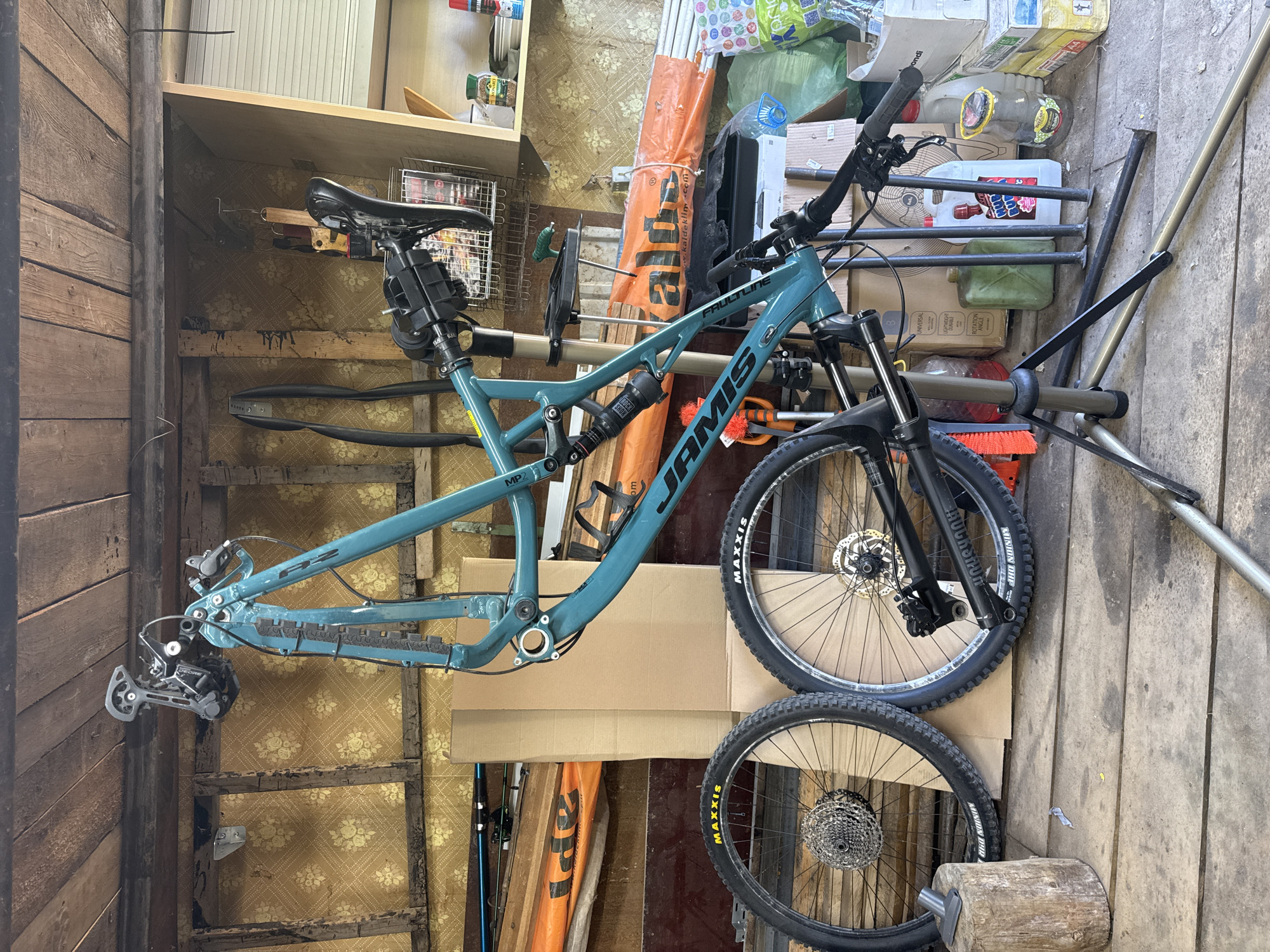A cyan JAMIS mountain bike frame with front and rear wheels is suspended inside a dilapidated garage or shed. The bike is partially assembled, with the frame, seat, handlebars, front fork, and rear derailleur visible. The front and rear wheels, both with MAXXIS tires, are also present but detached from the frame.
The background reveals a cluttered and disorganized interior, with wooden walls and shelves filled with various items, including boxes, bottles, tools, and other miscellaneous objects. The wallpaper is peeling and aged, adding to the overall state of disrepair.
Based on the OCR, "www.kaldeklim.com," and "ROTATION ANGLE," it's possible the tools are used for plumbing work.
Based on the OCR, it's possible this place is in Chaikovsky, Russia.