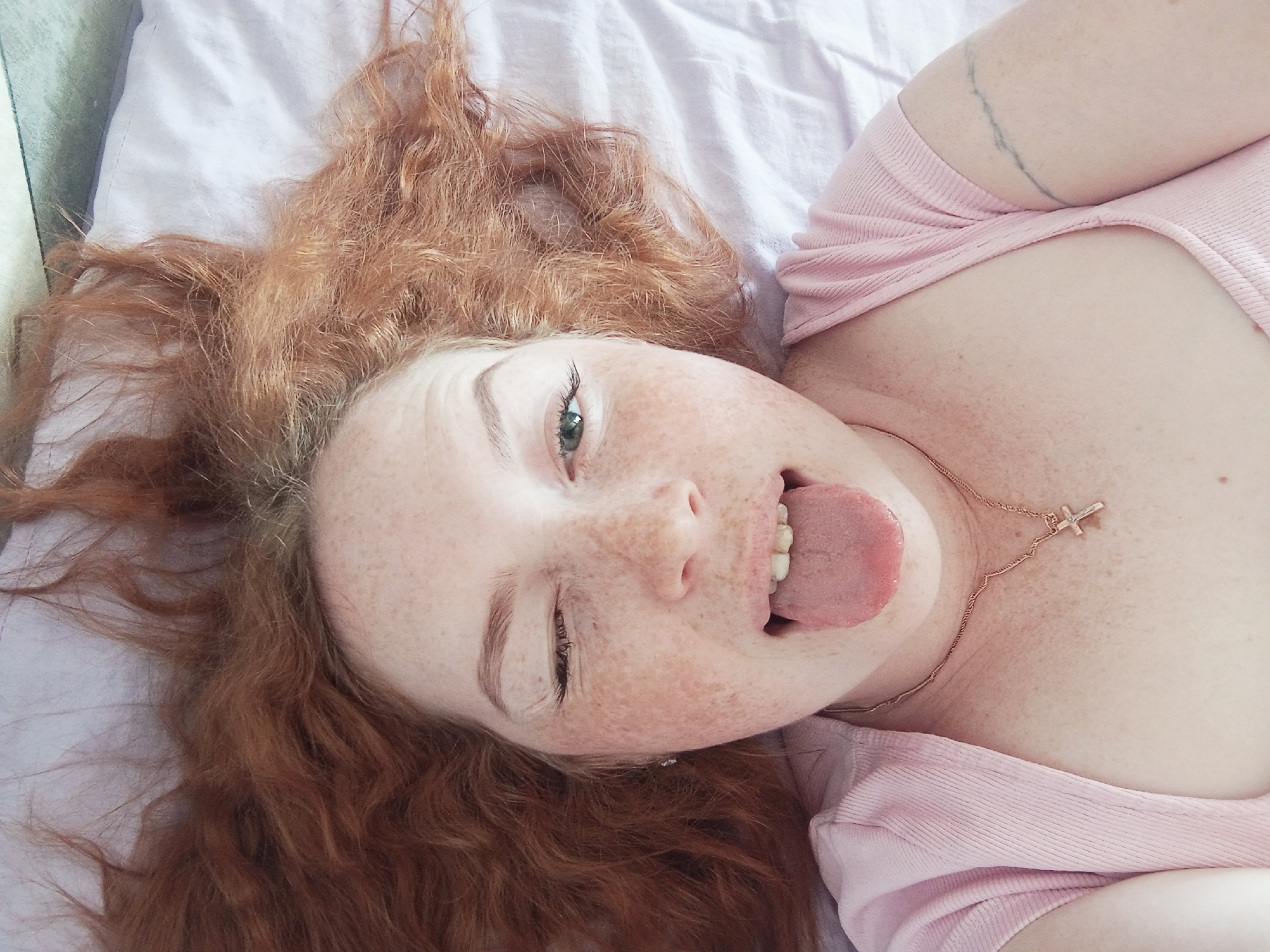 A pale-skinned woman with red, curly hair is lying on her back on a light purple bed sheet. She is winking her left eye and sticking her tongue out. Freckles are visible on her face and chest. She is wearing a pink shirt and a gold necklace with a cross pendant. A thin, linear tattoo is visible on her right arm. The image location is not discernible.