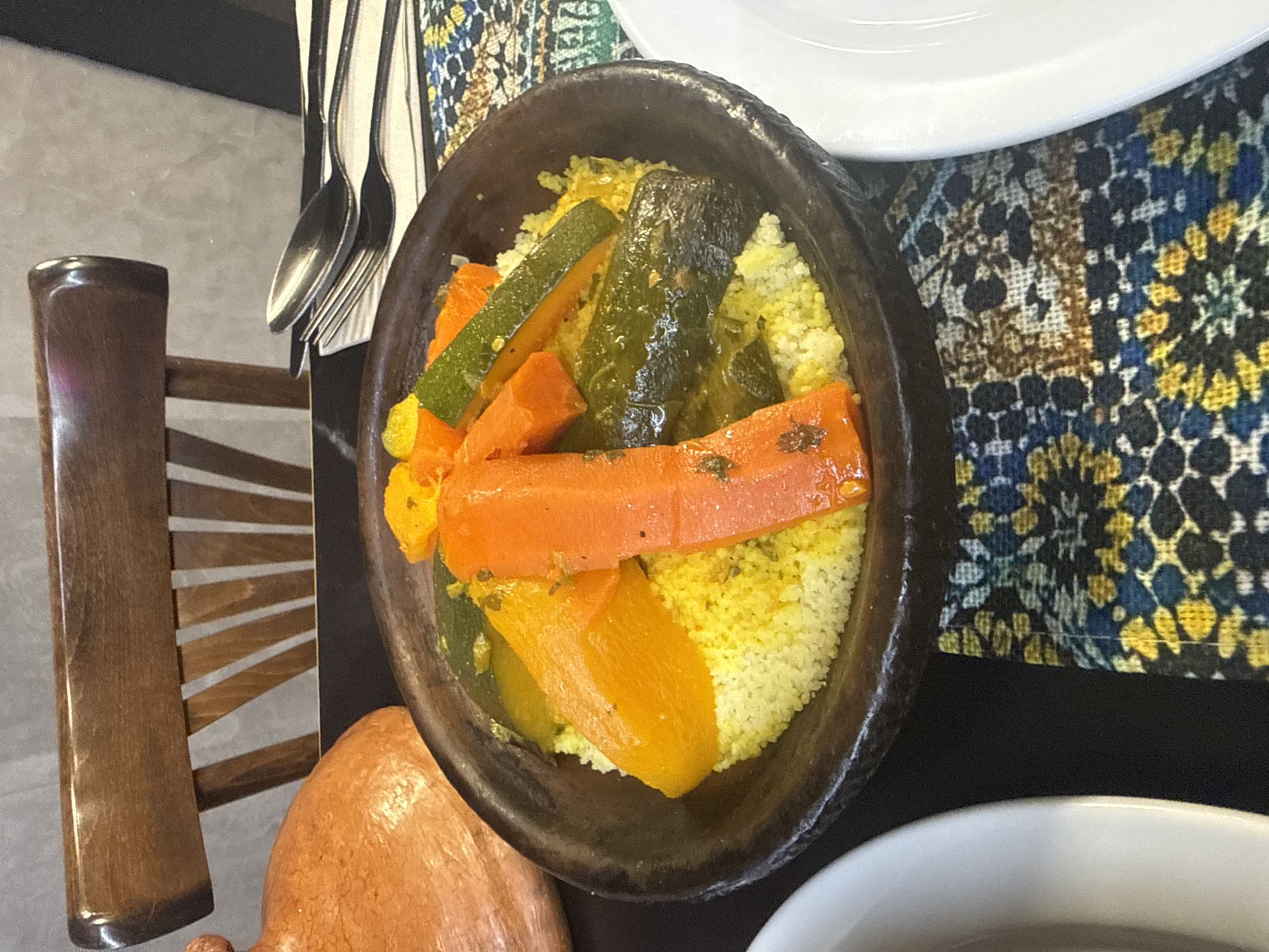 The image presents a top-down view of a meal, likely couscous, served in a dark earthenware bowl. The couscous is garnished with various vegetables, including zucchini and carrots, cut into large pieces. The bowl is positioned on a black surface next to a colorful patterned fabric. A set of silverware and a partially visible chair are also in the frame. The overall presentation suggests a dining setting in Barcelona, Spain, with the meal perhaps served in a restaurant or home known for its traditional cuisine.