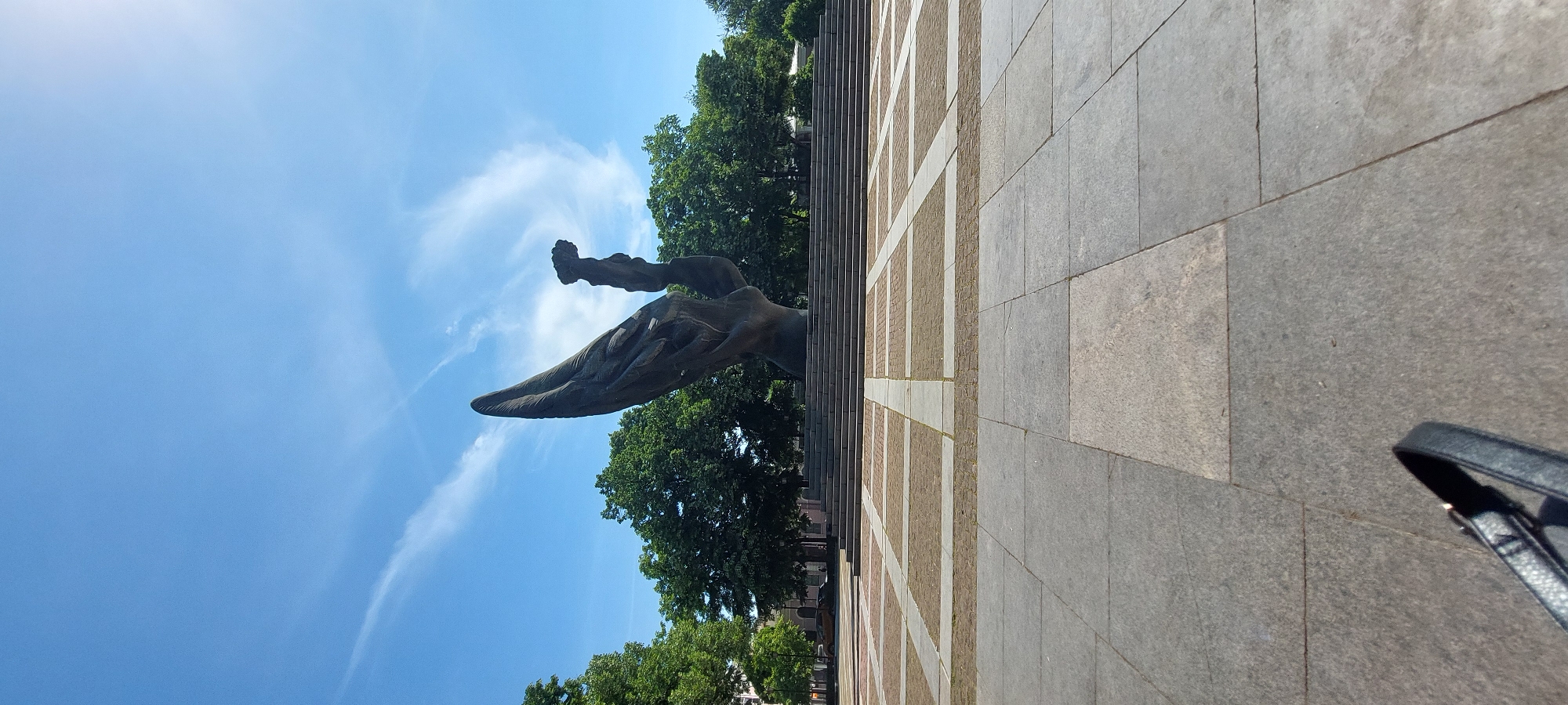 The image shows the Alyosha monument in Plovdiv, Bulgaria, viewed from a low angle. The bronze statue of a Soviet soldier with his hand raised towards the sky stands on a large stone base, surrounded by trees and a clear blue sky with wispy clouds. The paving stones in the foreground are grey. The corner of a black bag is visible in the lower right corner.