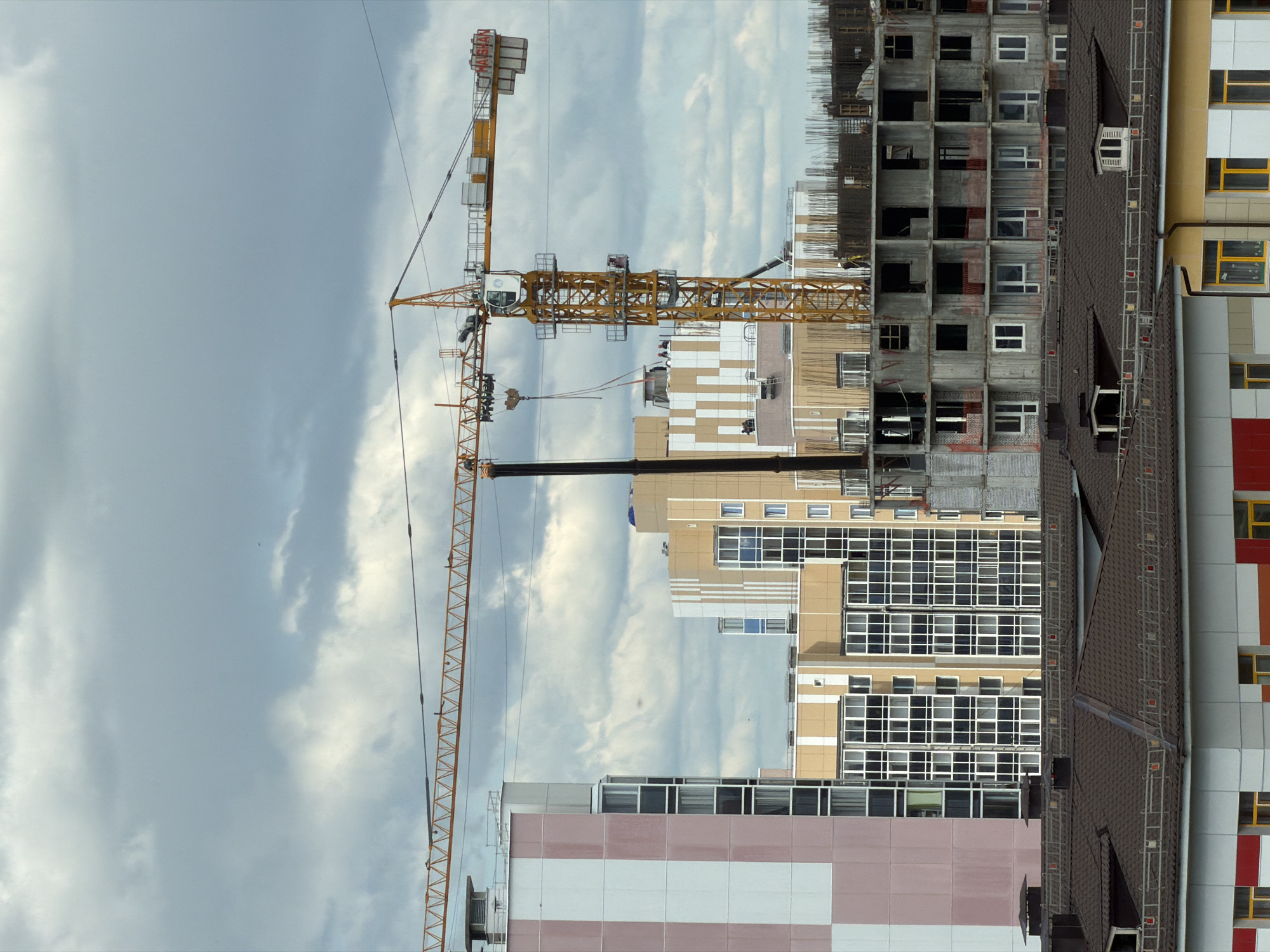This is an exterior, eye-level, full shot of a construction site in the city of Tsiolkovsky, Russia, on a cloudy day. In the foreground, there are two low-rise buildings with tiled roofs. The building on the left has a light gray facade with red and white horizontal stripes. The building on the right has a light yellow facade with white accents and yellow framed windows. Behind these, a large, multi-story building under construction is visible, with exposed concrete floors and rebar. Two tower cranes, one yellow and one dark gray, dominate the midground. The yellow crane has "HAISHAN" written on its boom. In the background, several tall, modern apartment buildings are visible, with pink and white, and beige and white facades, and many windows. The sky is heavily overcast with dark gray clouds. A few people are visible on the construction site, including one person in a hard hat on the yellow crane and several figures on the lower levels of the building under construction.