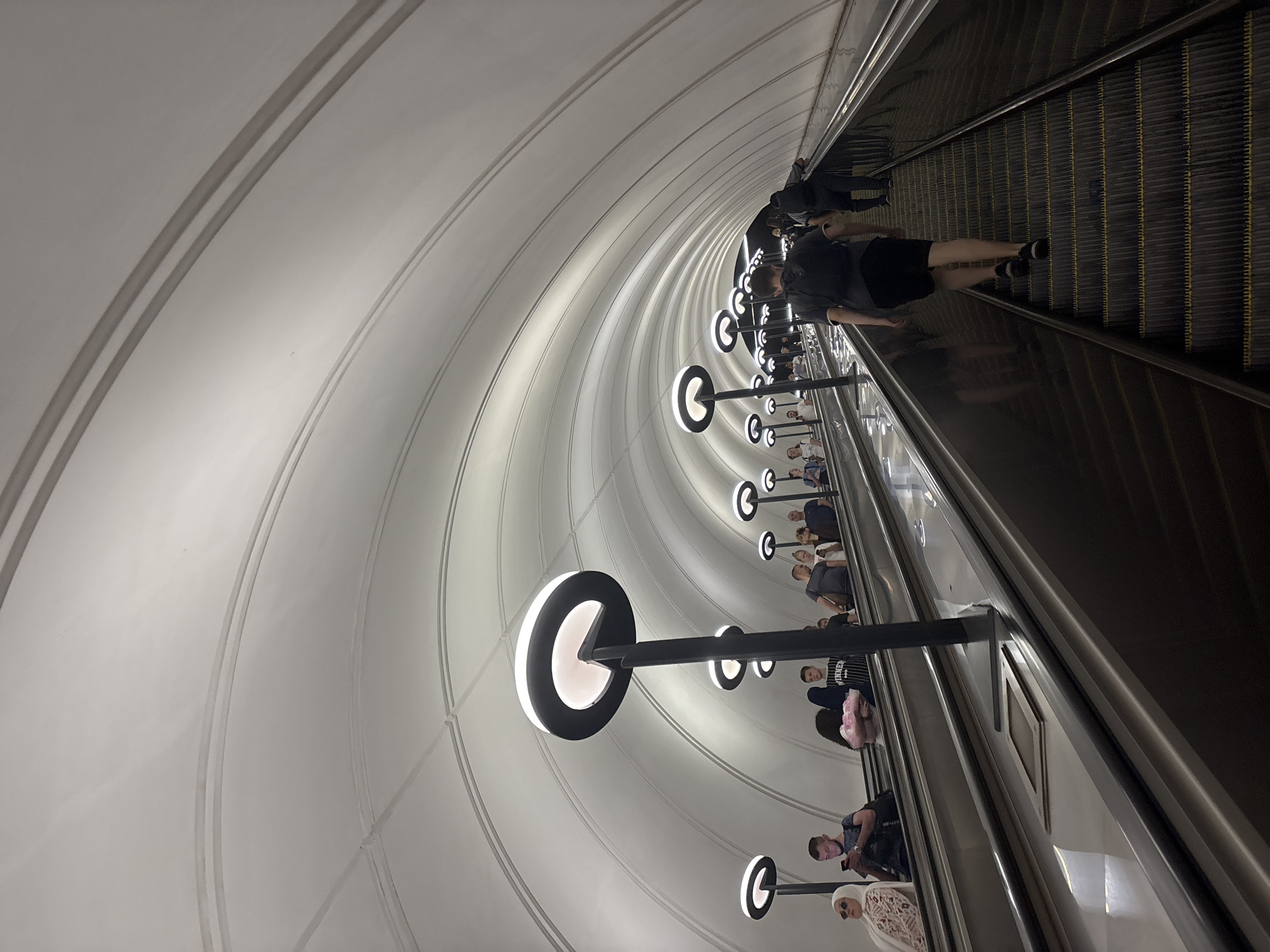 The image depicts a long, wide escalator in a subway station. The ceiling is a vaulted white structure with multiple arched supports. Circular lamps with white light are mounted on tall black poles, evenly spaced along both sides of the escalator. Several people are visible on the escalator, some ascending and others descending. The individuals are dressed in casual attire. The escalator itself is made of dark metal steps with yellow safety lines. In the foreground, a young man with a backpack is ascending the escalator. The scene suggests an active and functional public transportation hub. The location is identified as the city of Khimki, Russia.