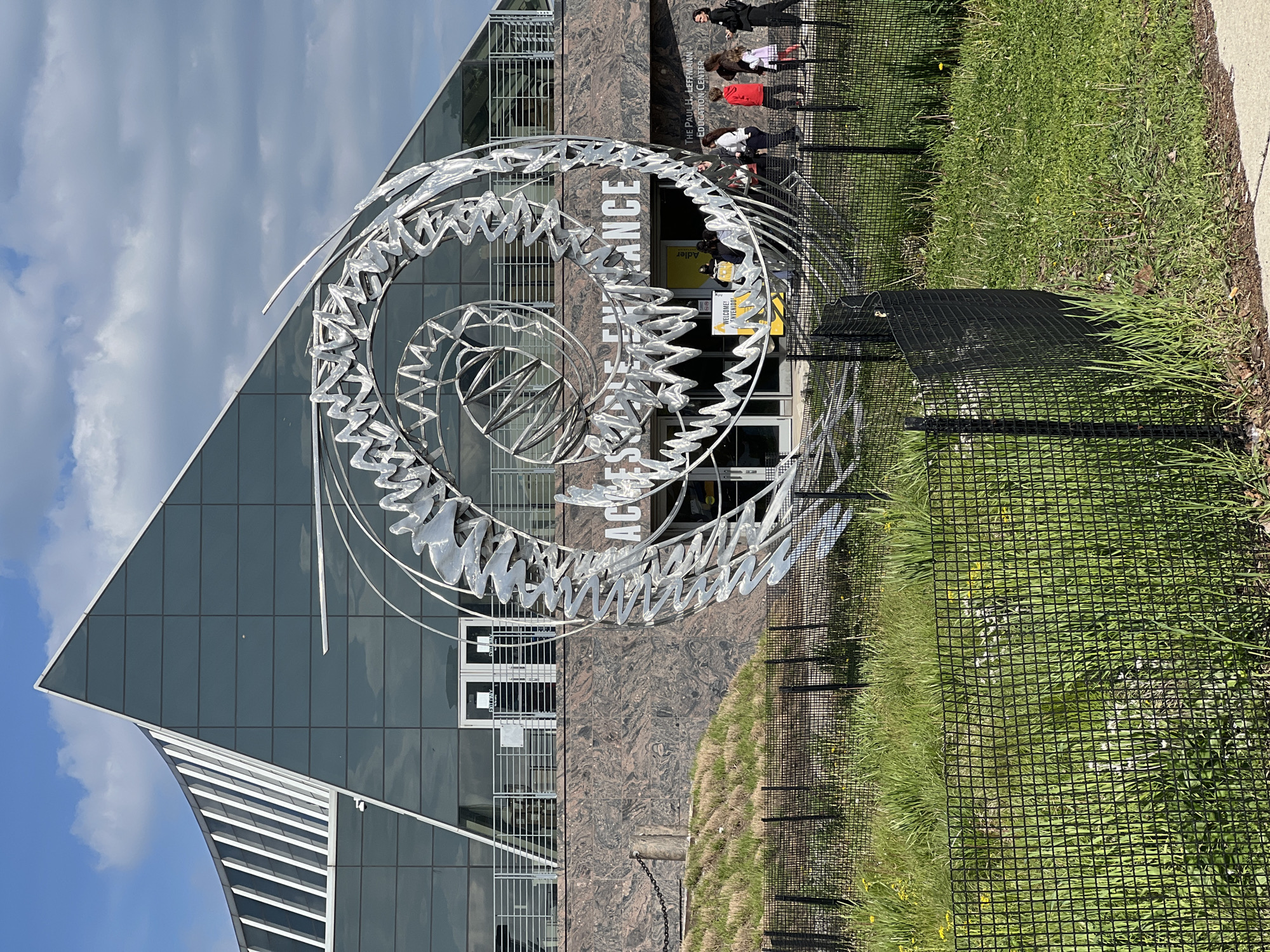 The image shows the Adler Planetarium in Chicago, United States. The planetarium's entrance is visible, marked by a large, silver, spiral-shaped sculpture. People are entering the building. A black mesh fence is located near the sculpture and grassy area. The building's exterior is characterized by geometric shapes, including a triangular glass facade. The sky is partially cloudy.