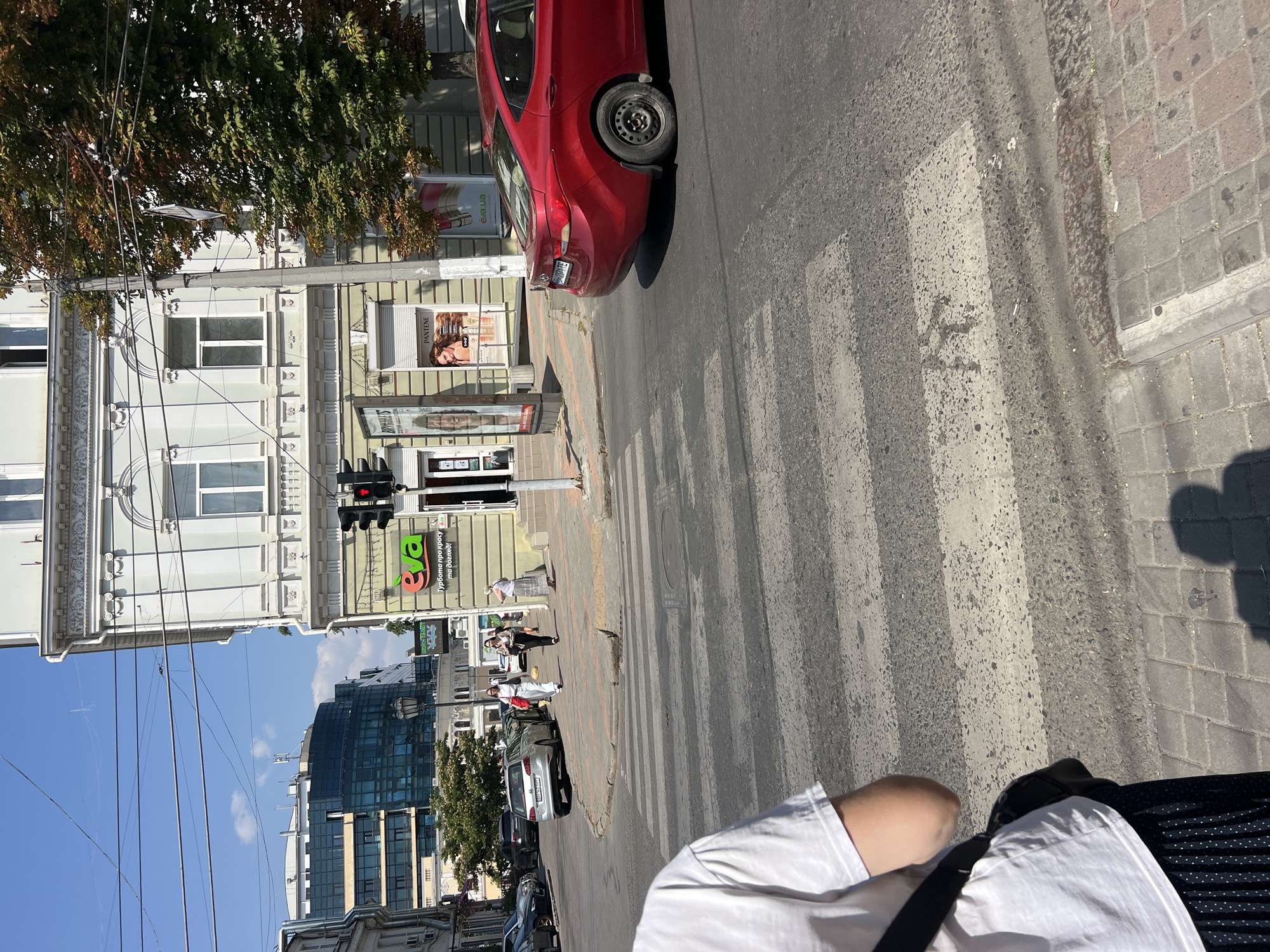 This is a street scene in Odesa, Ukraine, on a sunny day. In the foreground, a pedestrian is standing at a crosswalk, partially obscuring the view of the street. A red car is stopped at the crosswalk, waiting for pedestrians to cross. In the background, there is a multi-story building with an "eva" cosmetics store on the ground floor. Further back, a modern glass building can be seen. The street is paved with asphalt, and there are power lines running overhead. A traffic light is visible, currently red, indicating that vehicles should stop. Several other cars are parked or driving on the street, and a few pedestrians are walking on the sidewalk.