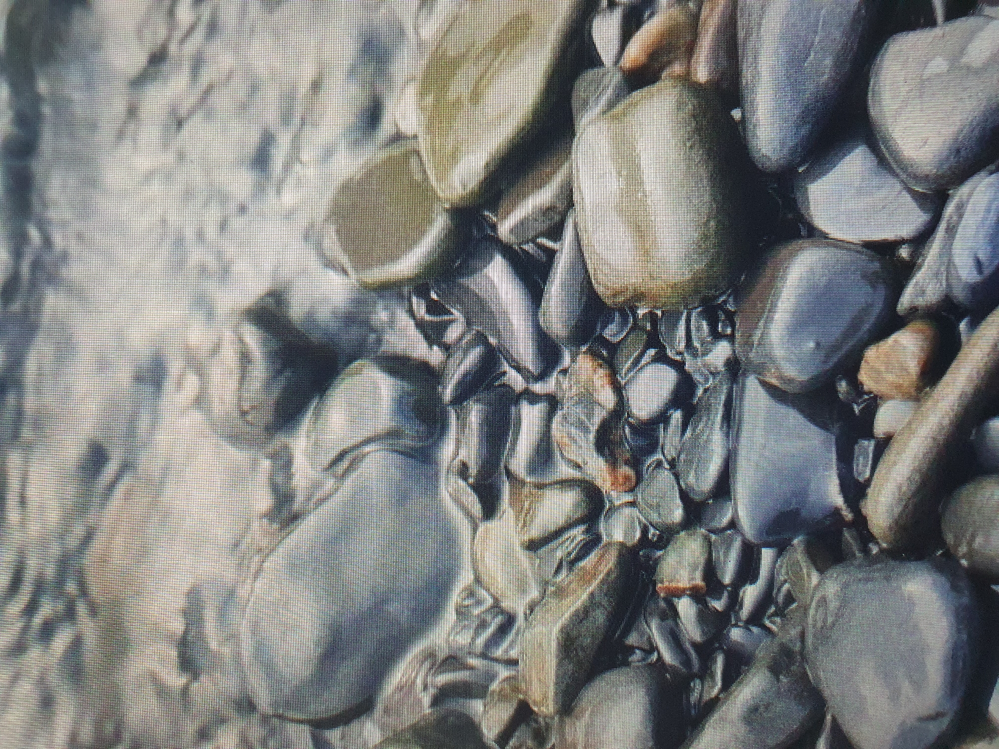 The image displays a close-up view of smooth, wet pebbles on a riverbed or shoreline. The stones are predominantly dark gray and blue, with some lighter, tan-colored stones interspersed. The surface of the pebbles appears polished and reflective, indicating they are submerged in or recently exposed to water. The water itself is visible in the upper portion of the image, appearing murky and reflecting the surrounding environment. The scene is focused on the textures and shapes of the stones, with no people or man-made objects present. The setting is natural, likely a body of water with a pebble-strewn bank. There is no information in the image to suggest a specific location within Tashkent, Uzbekistan.