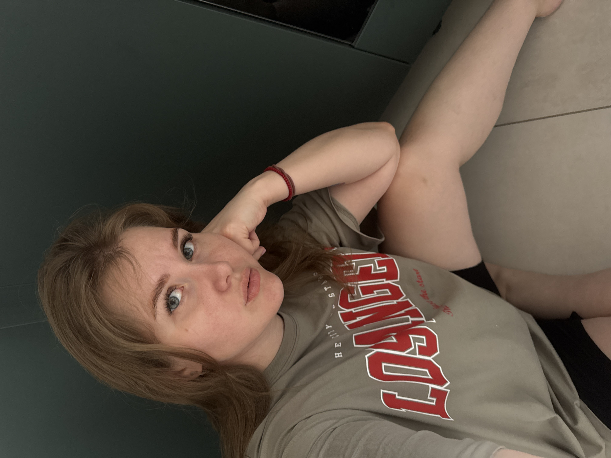 A woman is lying down, appearing to take a selfie. She has fair skin, light brown hair, and blue eyes. She is wearing a grey t-shirt with the word "LOSANGELES" printed on it in red and white. She also has a red bracelet on her left wrist. The background includes a dark grey wall and a light grey tiled floor. The location cannot be determined based on the image.