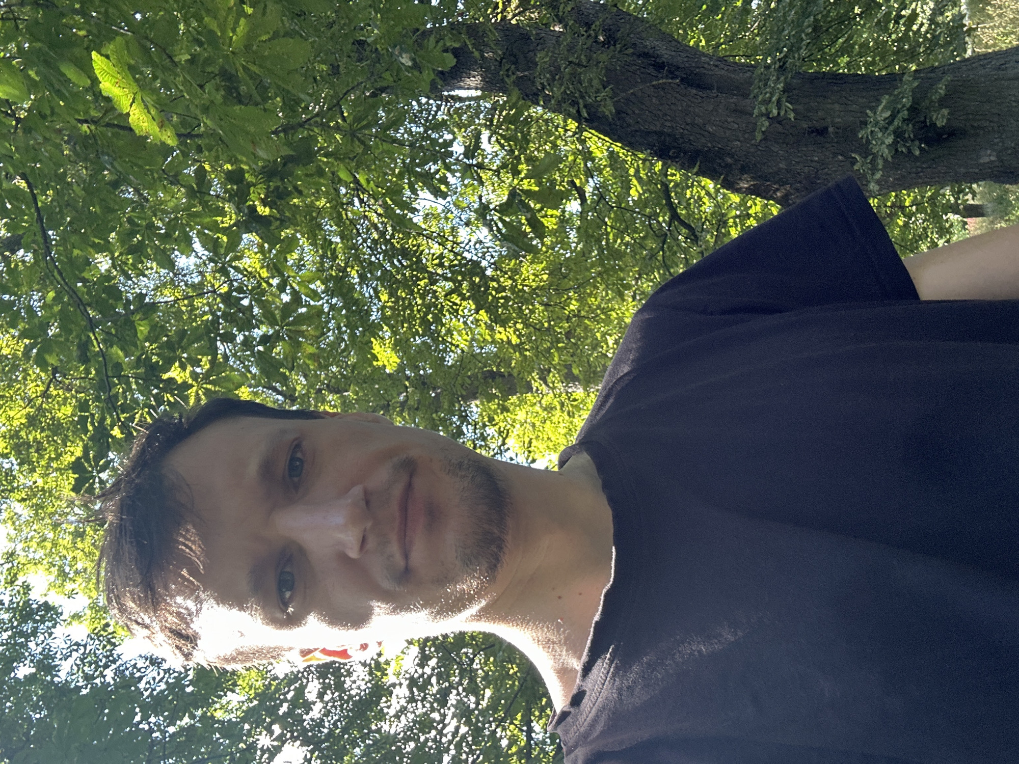 A man is taking a selfie outdoors with lush green trees and foliage in the background. He has short brown hair and is wearing a black t-shirt. The sunlight is filtering through the leaves, creating a dappled effect on his face and the surroundings. The location is identified as being in the city of Makiv, Ukraine. The man's expression is neutral, and he is looking directly at the camera. The overall scene suggests a casual, outdoor setting.