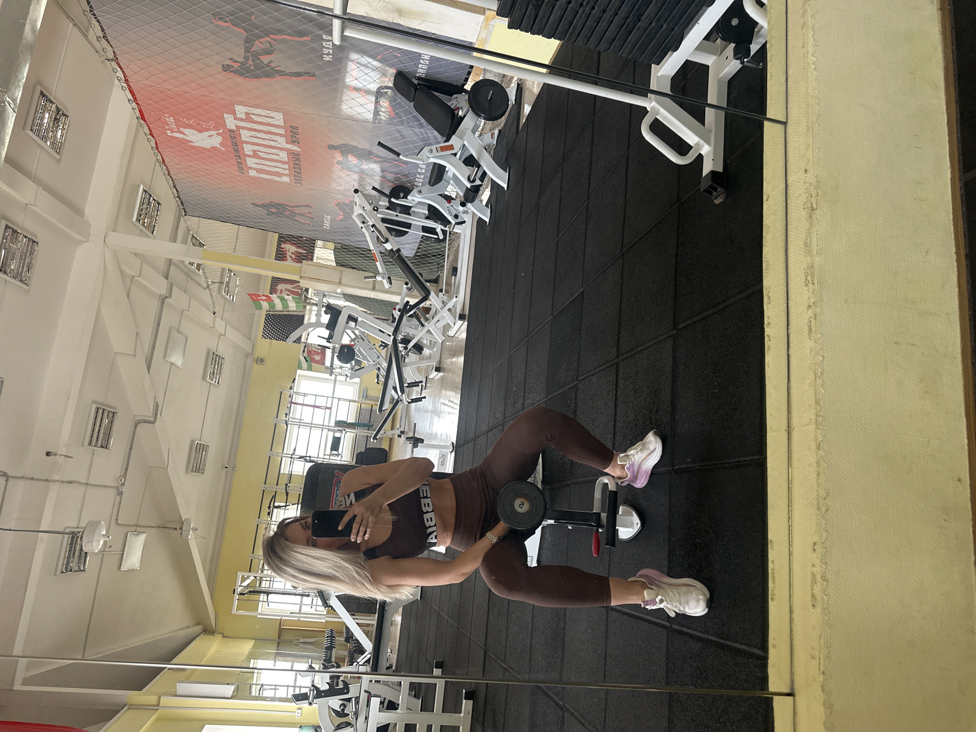 A blonde woman is taking a selfie in a gym that is reflected in a mirror. She is wearing a brown athletic top and leggings, and white running shoes with a lavender accent. She is holding a phone in her left hand and holding a dumbbell in her right hand. She is sitting on a weight bench. The gym has black rubber flooring and white walls with rectangular windows near the ceiling. There are various weight machines in the background. A large advertisement banner with a net over it is visible at the top. The location is likely in Chaikovsky, Russia, based on information in the image.