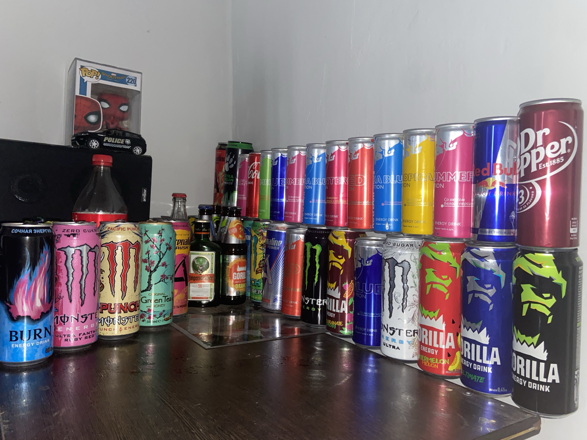 The media file displays a collection of energy drink cans, other beverages, and decorative items arranged on a wooden surface, likely a desk or shelf, in an indoor setting in Perm, Russia.
**Subjects:** The main subjects are numerous beverage cans, including a wide variety of Monster Energy drinks (e.g., Ultra Fantasy Ruby Red, Pacific Punch, Green Tea & Honey), Burn Energy, Red Bull (various flavors such as white peach, tropical fruits, nectarine, cranberry), Gorilla Energy Drink (watermelon melon, ultimate), Adrenaline Rush, and Adrenaline Power. Also present are a Dr. Pepper can, a Coca-Cola bottle, a Jägermeister bottle, and another smaller bottle. Above the beverages, there is a Funko Pop figure of Spider-Man in its box (labeled "POP! SPIDER-MAN 220") and a small black toy police car. A black electronic device, possibly a speaker, with "aceline" visible, sits behind the front row of drinks.
**Activity or event:** No active event is taking place; the image captures a static display or collection of items.
**Notable details:** The collection is diverse, featuring many different brands and flavors of energy drinks, some of which are stacked or arranged in rows, suggesting a collector's display. The lighting is artificial, indicating an indoor environment. Visible text on the cans includes various flavor descriptions in Russian, such as "СОЧНАЯ ЭНЕРГИЯ" (Juicy Energy) on the Burn can, "ZERO CUKR" on Monster cans, and "Бодрит тело и дух" (invigorates body and spirit) on some energy drinks. The Dr. Pepper can advertises "OF 23 FLAVORS." The Jägermeister bottle is marked "56".