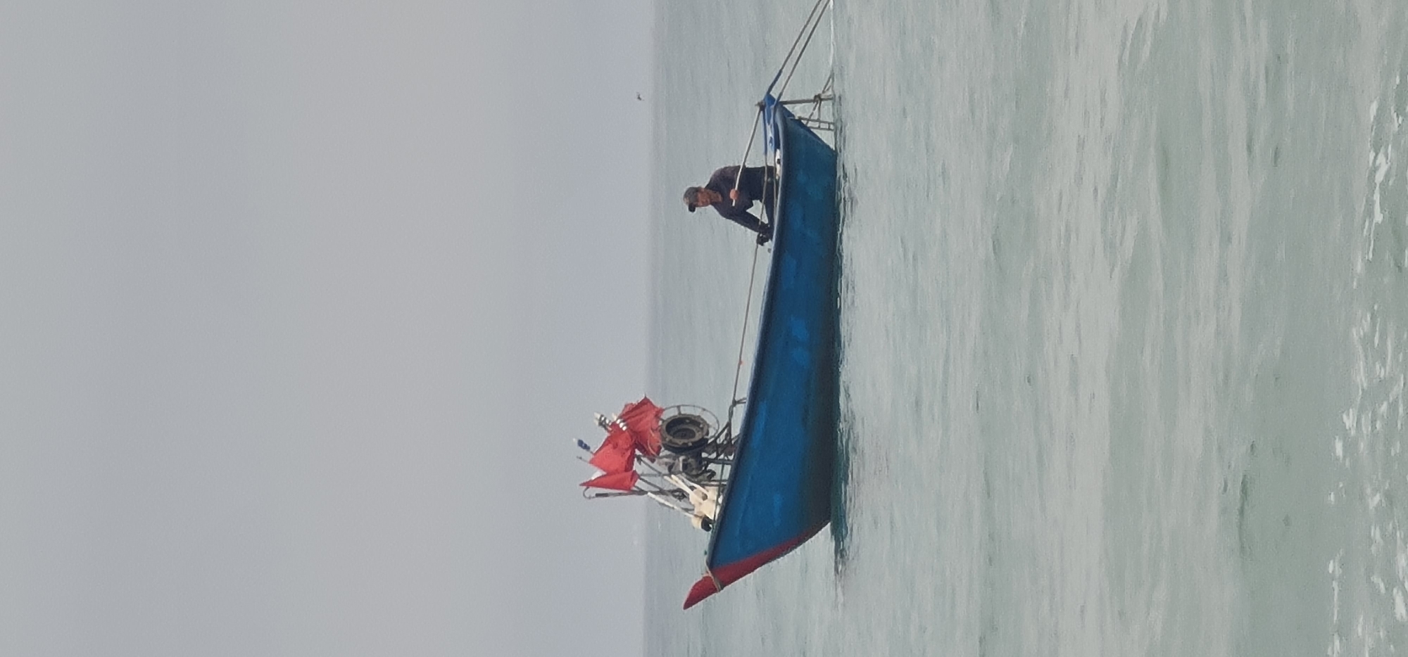 A medium shot depicts a fisherman on a small, blue boat in the water, likely off the coast of Đà Nẵng, Vietnam. The boat, painted red at the front, is equipped with a red flag and a fishing gear mechanism. The man is seen bent over near the front of the boat. The water is a light gray color, and the sky is overcast, creating a monochromatic palette.