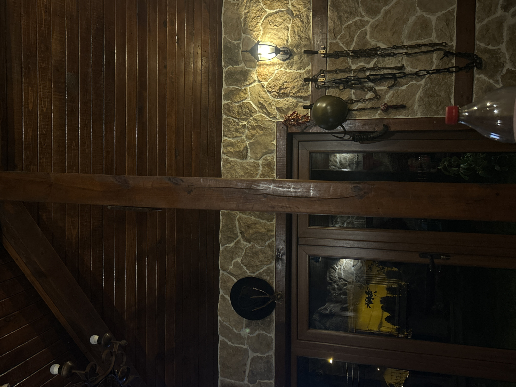 The image depicts the interior of a building, likely a restaurant or pub, in Mirovyane, Bulgaria. The wall is constructed with a combination of wooden paneling and stonework. Various decorative objects are mounted on the wall, including chains, a medieval-style helmet, and a black hat with antler-like protrusions. A wall-mounted light fixture is also visible. A thick wooden beam runs horizontally across the image. A large window with a wooden frame is present, showing reflections of the outside environment. The lower left corner of the image shows a portion of a decorative chandelier. A plastic bottle partially obscures the upper right corner of the image.
