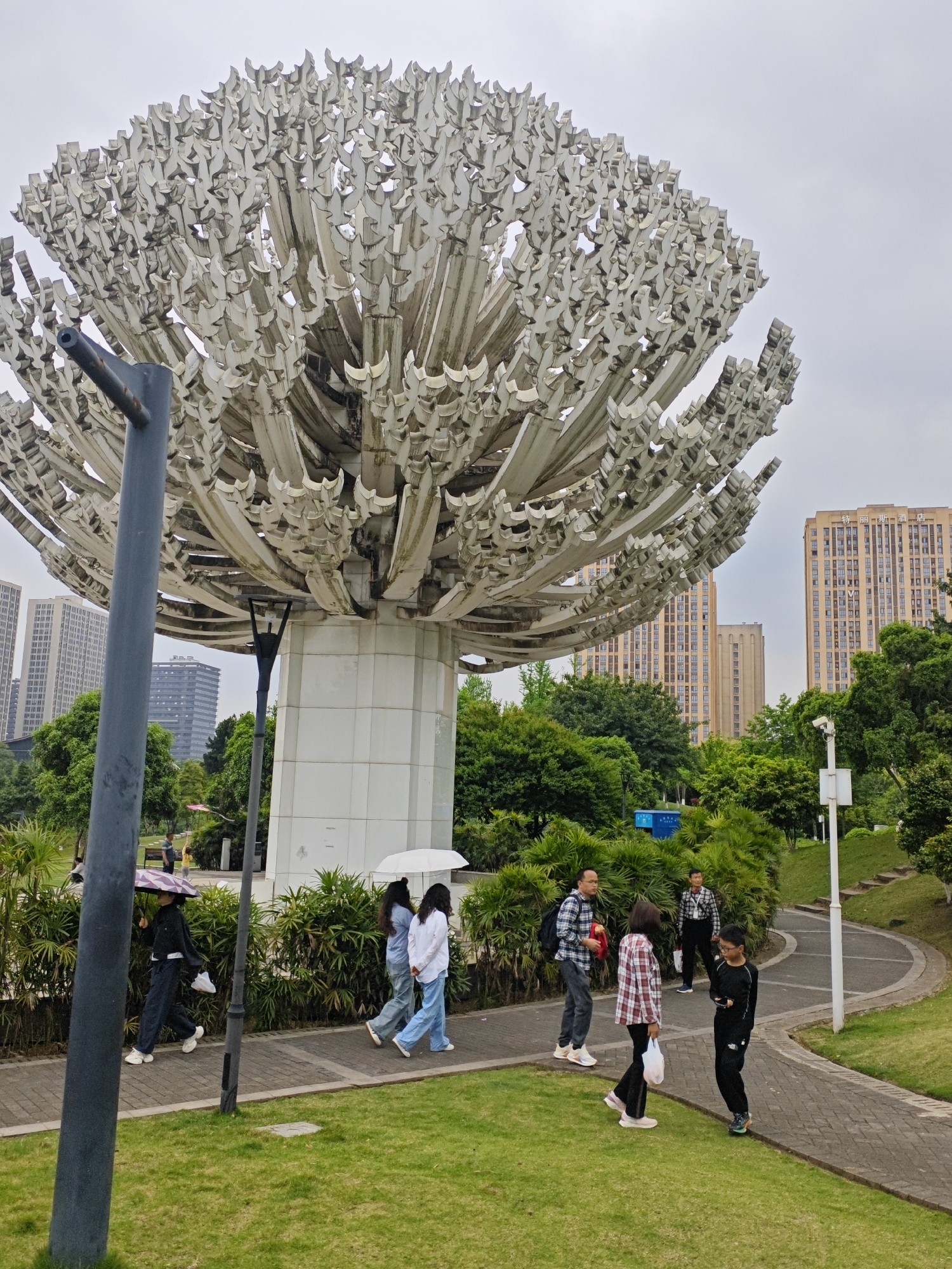 The image captures a scene in what appears to be a public park, situated in the Chongqing High-tech Zone, China. Dominating the foreground is a large, abstract sculpture resembling a tree, constructed from white metal or composite materials. The sculpture is positioned on a tiled base, and several people are seen walking along a paved pathway in the park.
To the left, an individual carries a purple umbrella. Two other figures walk in the same direction, one carrying a white umbrella. Further along the path, a group of three people is present, one holding a red object. A solitary person walks ahead of them, looking down.
The background consists of lush greenery, including trees and shrubbery. Tall buildings are visible in the distance, indicative of an urban setting. A lamp post and surveillance camera are also present in the park. The sky is overcast, creating a diffused natural light.