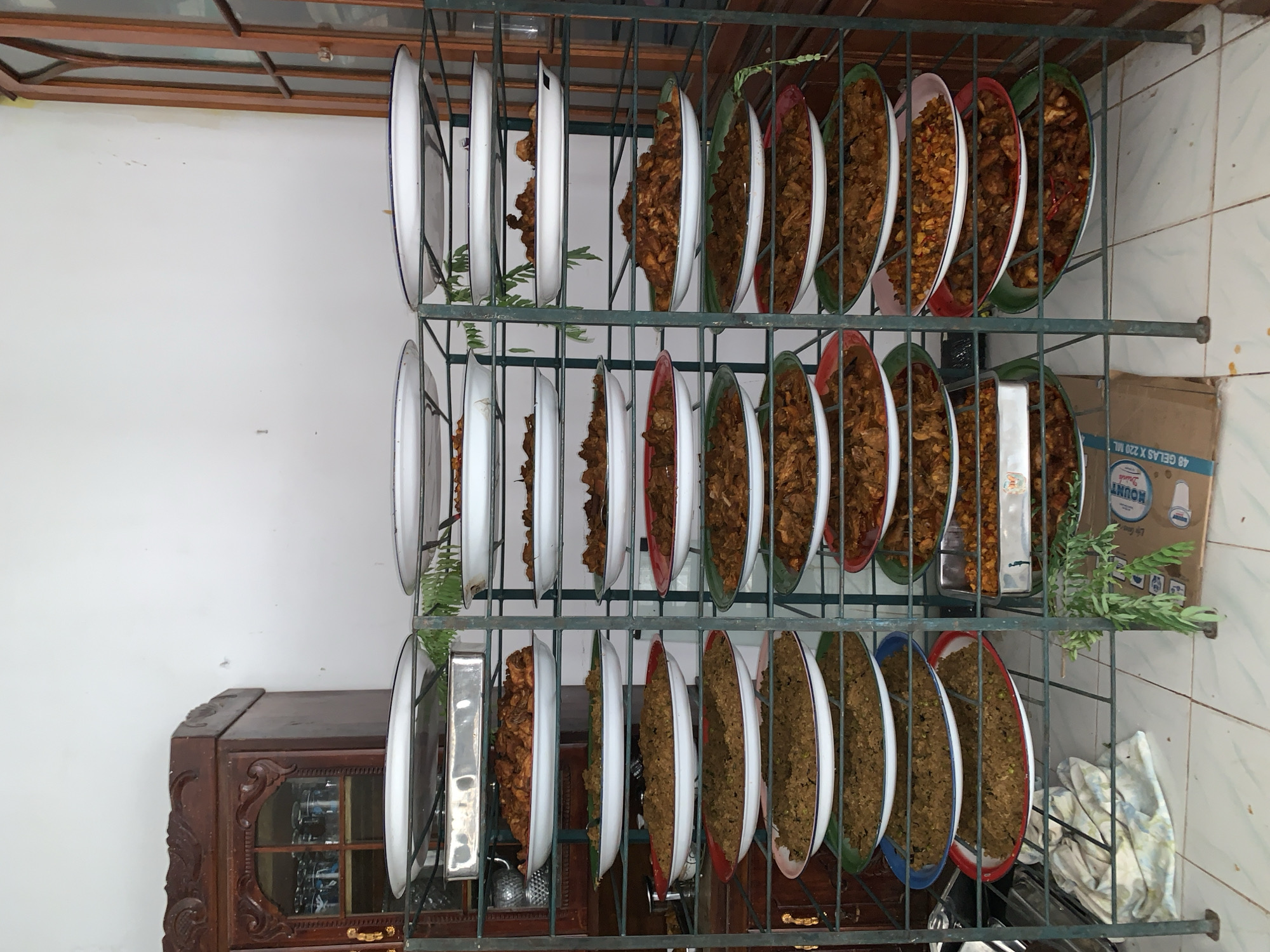 The image depicts a rack of numerous plates filled with food, likely in a food stall or restaurant setting, potentially in Uleepulo, Indonesia, given the style of the food and the cabinetry. The rack is made of dark metal and has multiple shelves, each holding several oval-shaped white enamel plates with colored rims. The plates are stacked in two columns on each shelf. The food appears to be a variety of Indonesian dishes, including rice, curries, and stir-fried items, with colors ranging from light brown to dark red. Some plates have green rims, others red, and some blue. In the background, a dark wooden cabinet with glass doors, possibly a display case or storage, is visible to the left. A similar tall wooden structure with glass doors is on the right side of the image. The floor is tiled with light-colored squares. Some green foliage, possibly decorative or a plant, hangs over some of the shelves. A cardboard box with printing, partially visible at the bottom right, suggests a commercial area. The overall impression is of a large quantity of prepared food ready for serving.