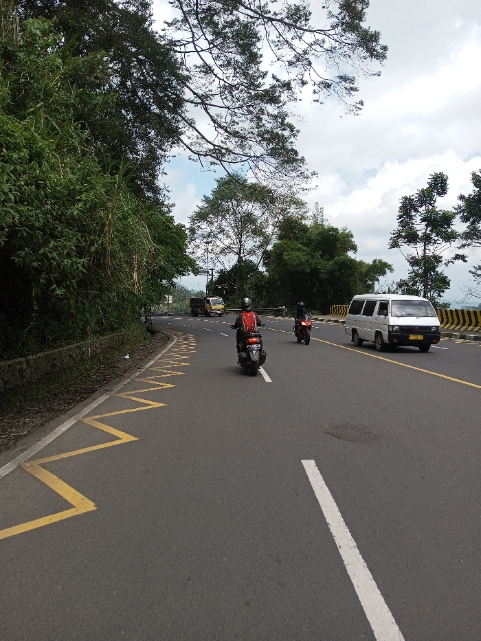 This image depicts a multi-lane asphalt road curving gently to the right, under a partially cloudy sky, located in the city of Boommlog, Bhutan. In the foreground, a dark-colored scooter is ridden by an individual wearing a red jacket and a helmet, moving away from the camera. Immediately behind this scooter, a red motorcycle is operated by another helmeted individual in dark attire, also moving away.
The road features a solid white line extending diagonally across the lower right foreground, which serves as a lane divider. On the left shoulder, yellow zigzag markings are painted on the asphalt, bordered by a solid white line and an adjacent concrete drainage ditch with dense green vegetation on a raised bank. In the opposing lane, a white van, appearing to be a Mitsubishi L300 model, is driving towards the camera. Further down the road in the opposing lane, a yellow truck is visible. A black and yellow striped barrier runs along the right side of the road, separating it from a vegetated slope. The background consists of a mix of broadleaf and conifer trees, with a utility pole or traffic signal structure visible in the distance.