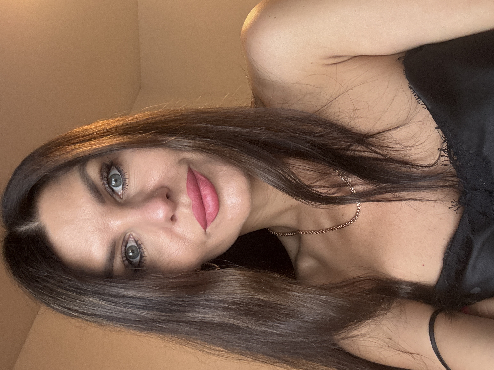 The image is a close-up selfie of a woman with long, dark brown hair, fair skin, and blue-green eyes. She is wearing a black top with lace trim and a delicate gold chain necklace. Her makeup includes pink lipstick and mascara. The background is a blurred, light brown wall, suggesting an indoor setting. The lighting is warm, with a golden hue. The image does not provide any information about the location within the city of Chaikovsky, Russia.