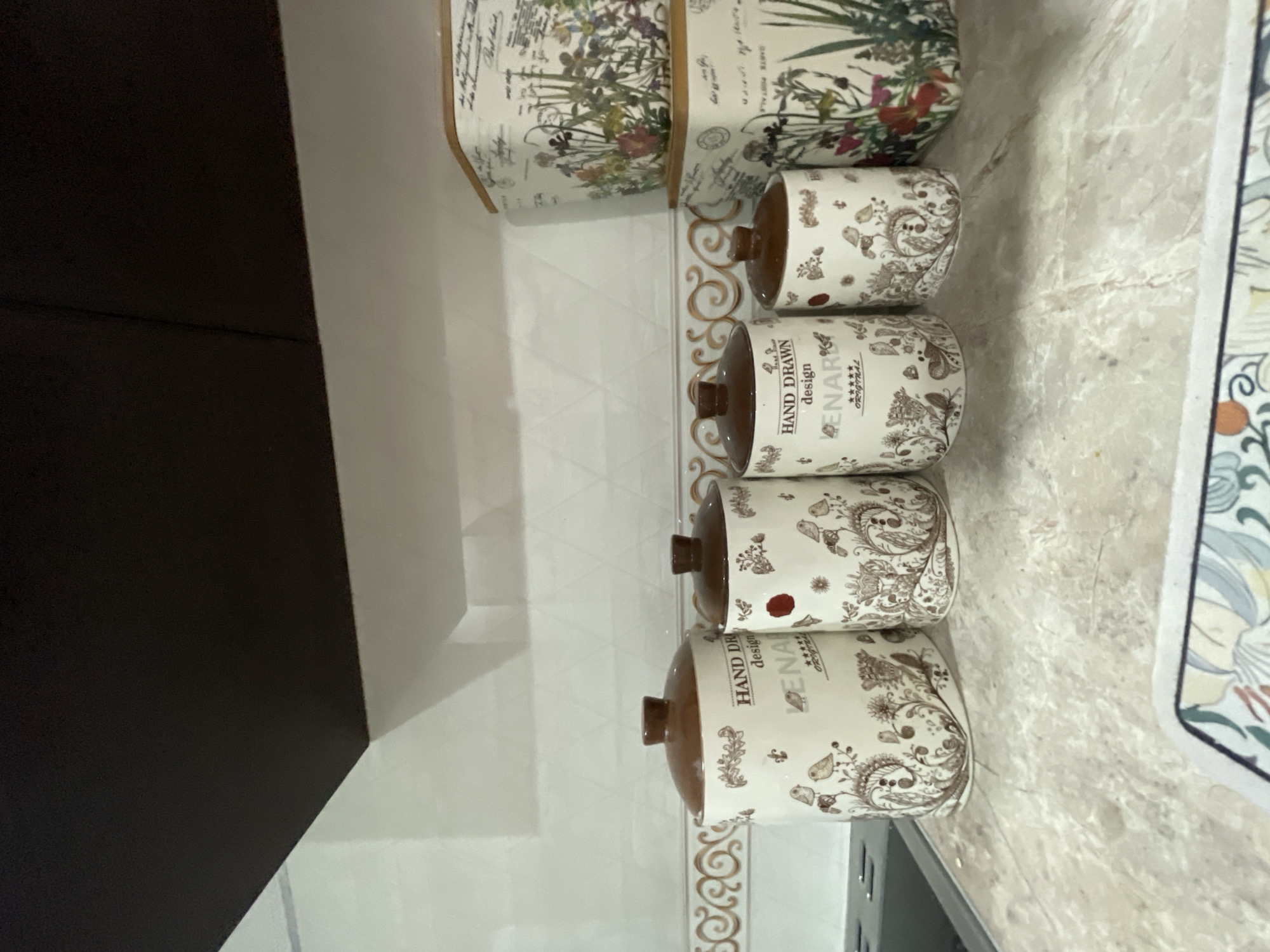The image shows a close-up of four decorative ceramic canisters with brown lids and a hand-drawn design featuring birds and floral motifs, arranged on a speckled countertop. To the right of the canisters, two larger decorative boxes with floral and script designs are stacked. The background consists of a tiled wall with a patterned border. The scene appears to be in a kitchen or pantry setting. There are no people or specific actions depicted. The image does not contain any information that would allow for the identification of its location in Ulan-Ude, Russia.
