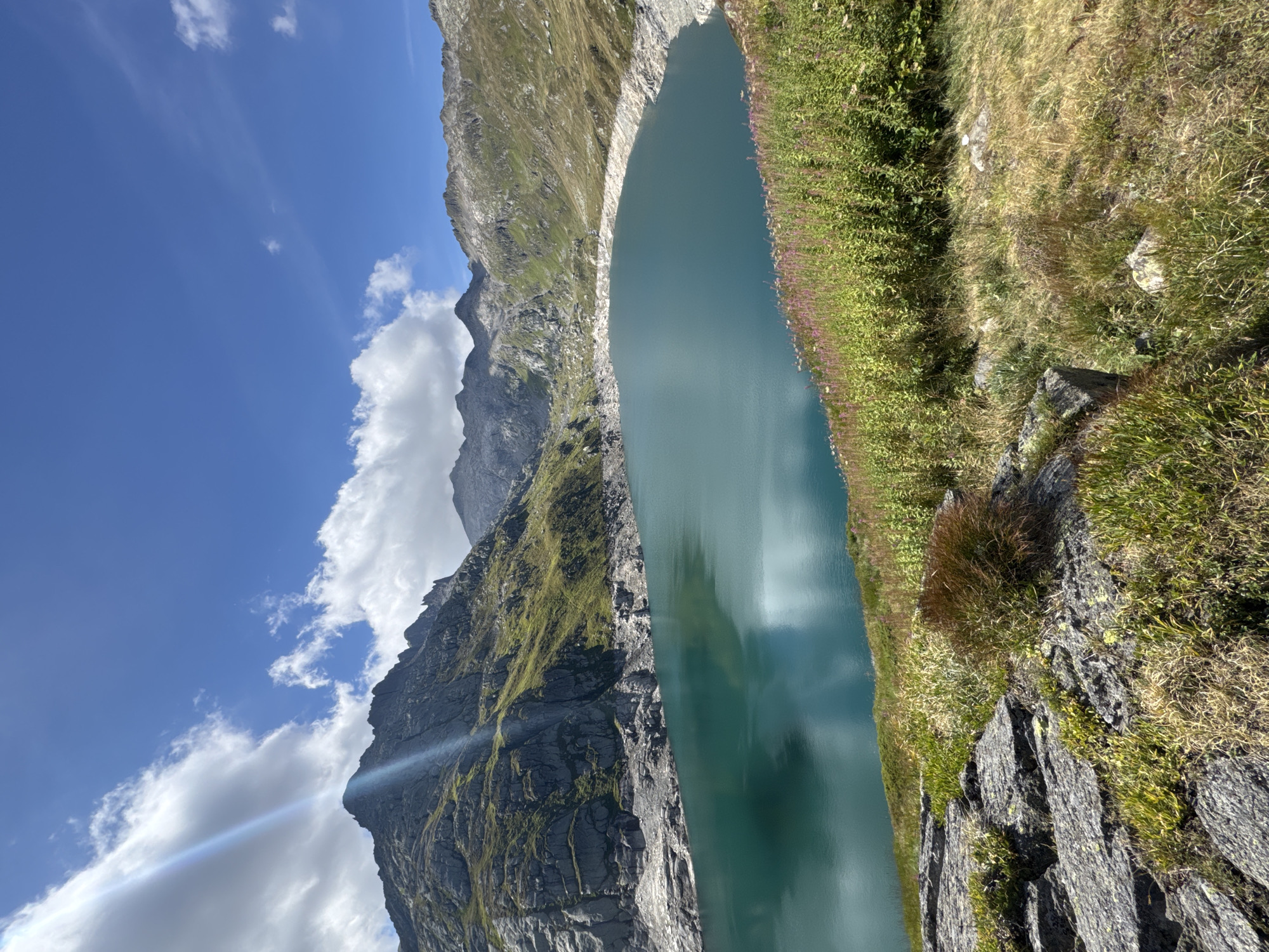 The image shows a scenic view of a glacial lake in a mountainous region, likely in the vicinity of Airolo, Switzerland. The turquoise lake is surrounded by rugged, rocky mountains covered in patches of green vegetation. The sky is a clear blue with scattered white clouds, and a sunbeam cuts across the frame, illuminating a section of the mountain. The foreground consists of rocks and grass, with a dense patch of flowering plants adding a touch of color along the edge of the lake. The overall impression is one of natural beauty and tranquility.