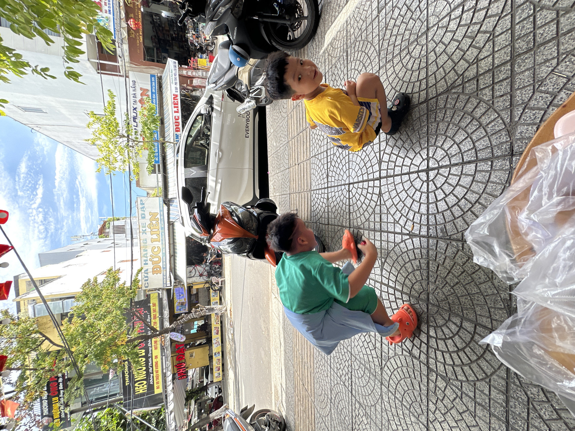 This image depicts an urban street scene in Đà Nẵng, Vietnam, specifically near 52 Trần Kế Xương street, identified by shop signs visible in the background. Two young boys are actively engaged on a patterned grey sidewalk in the foreground.
The boy on the left wears a green short-sleeved shirt, light blue shorts or a skirt-like garment over shorts, and orange sandals shaped like animal heads. He is bending forward, looking down, and appears to be interacting with a small orange object on the pavement with his right hand. The boy on the right, wearing a yellow t-shirt with "VARSITY" printed on the back, patterned shorts, and dark sandals, is in a squatting position, looking directly towards the camera with a slight smile. In the immediate foreground, out of focus, are several clear plastic bags containing light-colored items, possibly food, resting in a brown container.
The background reveals a bustling street environment. Various motorcycles and scooters are parked along the curb, including an orange and black scooter behind the boy on the left, and a black Honda scooter behind the boy on the right. A white SUV, possibly a VinFast Lux SA2.0 with "EVERYBODY" printed on its side, is parked further down the street. Buildings line both sides of the street. On the left, a multi-story building displays a large black sign for "THƯỢNG HEO NƯỚNG LŨ ONG ĐÀ NẴNG" (Grilled Pork Lu Ong Da Nang) and a smaller yellow sign for "PHÚ ĐẠT." On the right, a shop named "CỬA HÀNG XE ĐẠP ĐỨC LIÊN" (Duc Lien Bicycle Shop) is visible, with its address "52 TRẦN KẾ XƯƠNG - TP ĐÀ NẴNG" and phone numbers clearly displayed. Green trees with sparse foliage line the street, and red decorative flags are strung across overhead. The sky above is blue with scattered white clouds.