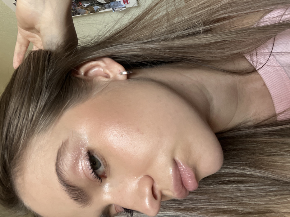 A close-up, indoor portrait features a young adult female, viewed from the left profile. Her fair skin displays a radiant complexion with subtle pink blush on the cheekbones and light pink, glossy lips. The left eye shows shimmering pink eyeshadow on the lid and long, dark eyelashes; her eyebrows are well-defined. Her long, straight hair, appearing light brown with lighter streaks, flows around her face and shoulders. A small, silver-colored hoop earring is present in her left earlobe. The subject's right hand is visible in the upper right quadrant, with fingers lightly touching her hair near the temple. She is wearing a light pink crew-neck top, partially visible at the bottom right. The background is out of focus, consisting of a plain, light-colored wall or surface with indistinct photographic elements faintly visible in the far distance. The specific city location of Saint Petersburg, Russia, cannot be determined from the visual information provided within the image.
