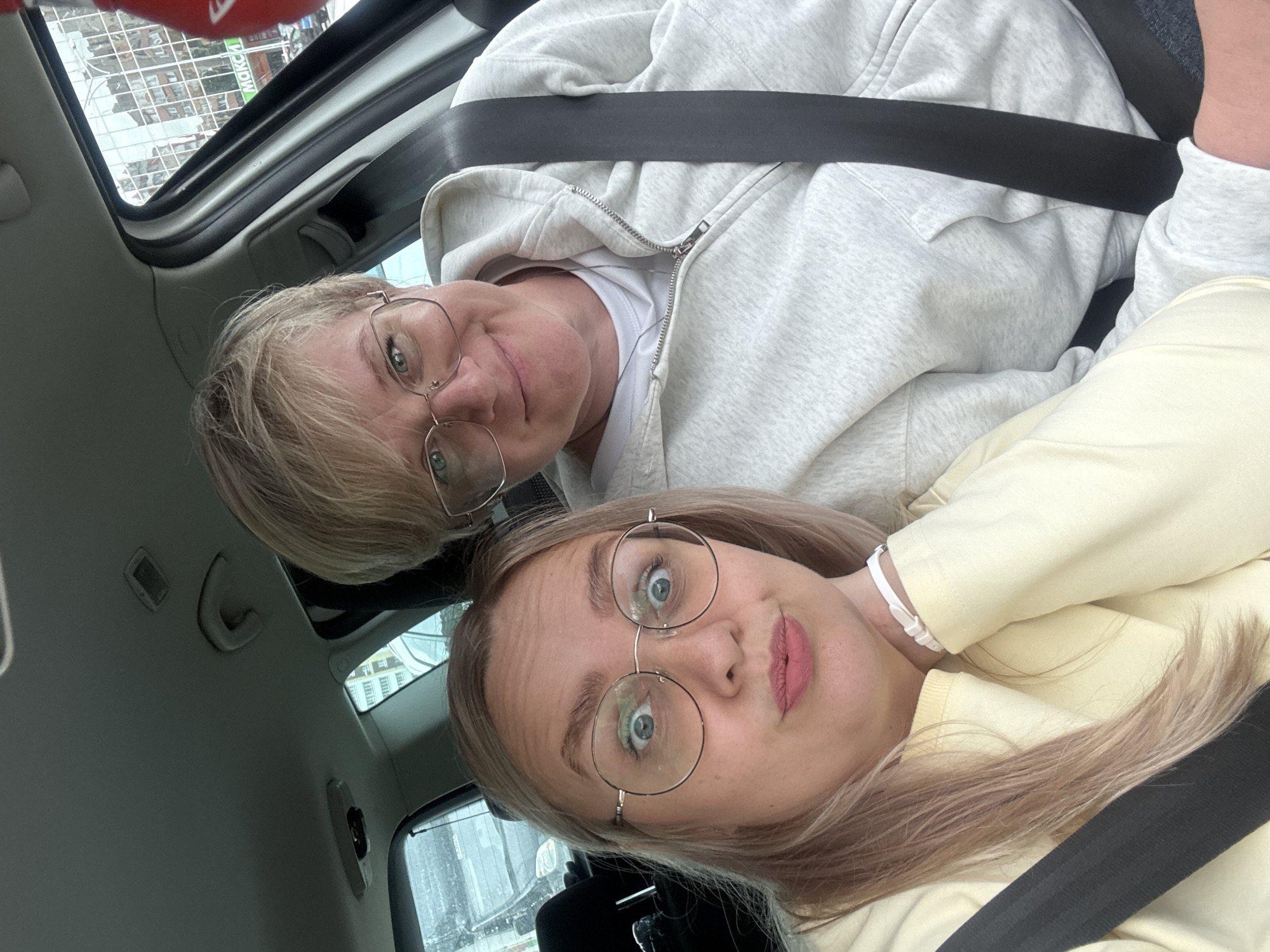 Two individuals are captured in a selfie taken inside a vehicle. The person in the foreground, on the left, has blonde hair and is wearing round glasses and a light yellow sweatshirt. They are looking directly at the camera with a surprised expression, their lips pursed. A seatbelt is visible across their chest. To the right and slightly behind is another person with short blonde hair, also wearing glasses and a light grey zippered hoodie over a white t-shirt, with a seatbelt across them. They are smiling and looking towards the camera. The background visible through the car windows suggests an urban environment with buildings. The overall lighting indicates it is daytime, though possibly overcast.