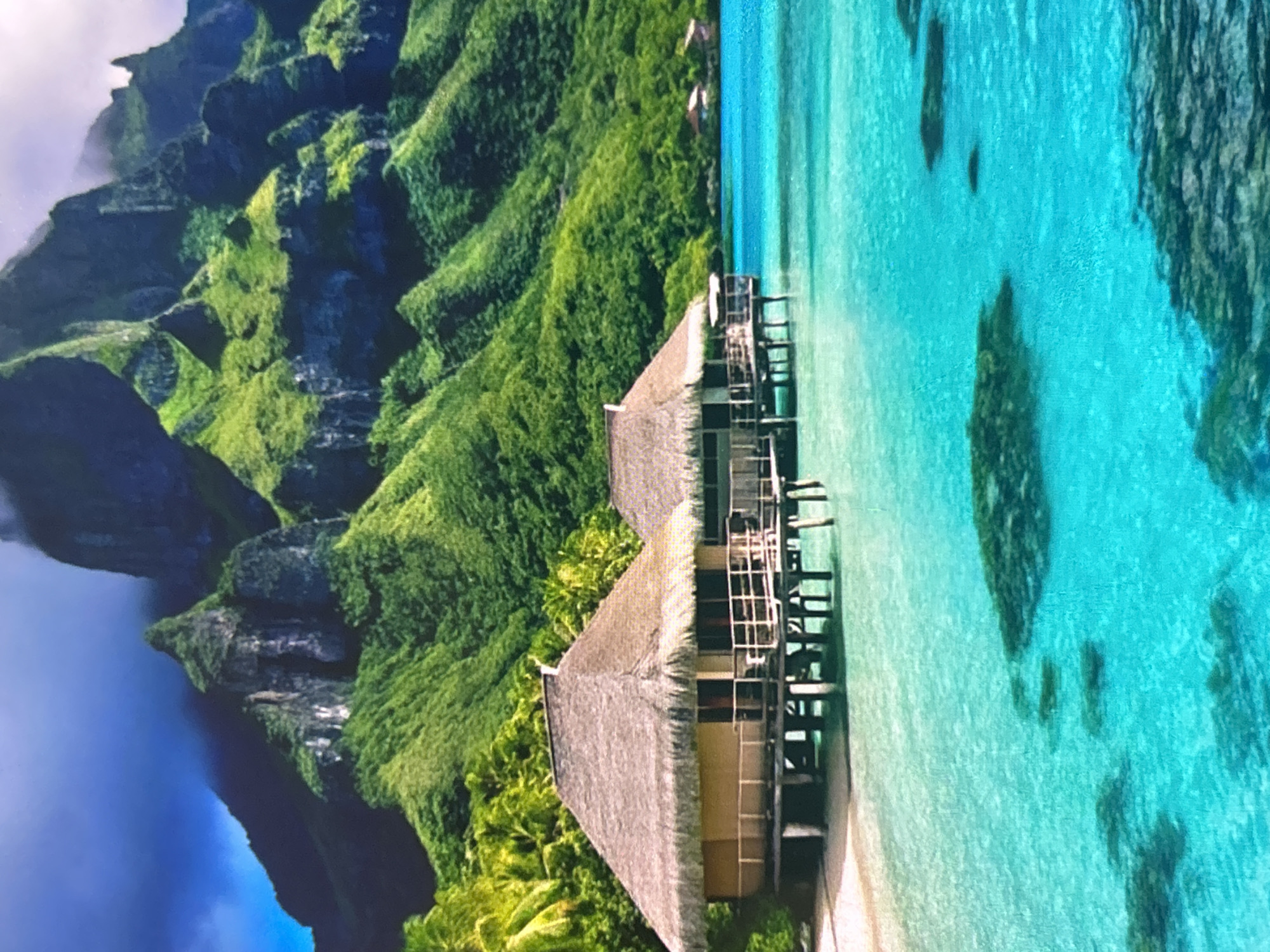 The image displays a vibrant tropical landscape featuring lush green mountains, crystal-clear turquoise waters, and overwater bungalows. The scene is oriented vertically in the provided image, but depicts a horizontal view.
On the left side of the image, majestic, steep mountains are covered in dense, verdant foliage, showcasing various shades of green. Rocky cliffs and outcrops are visible amidst the greenery, and some atmospheric mist or clouds cling to the higher peaks, suggesting a humid environment.
To the right, the brilliant turquoise and aquamarine ocean stretches out, revealing a clear sandy bottom with darker patches that appear to be coral formations or submerged rocks. A narrow strip of white sandy beach is visible along the shoreline. Two prominent overwater bungalows with thatched roofs stand on stilts over the water. These structures are connected by wooden walkways, suggesting a resort setting. Tropical vegetation, including palm trees, surrounds the bungalows and lines the edge of the land.
The scene depicts a peaceful and idyllic daytime setting, likely under sunny conditions, though the distant mountain peaks are veiled in light cloud. There are no people visible, and no specific activities are taking place, reinforcing the serene atmosphere. No text is visible in the image. The overall impression is one of a luxurious and pristine natural paradise, reminiscent of destinations like the South Pacific islands.