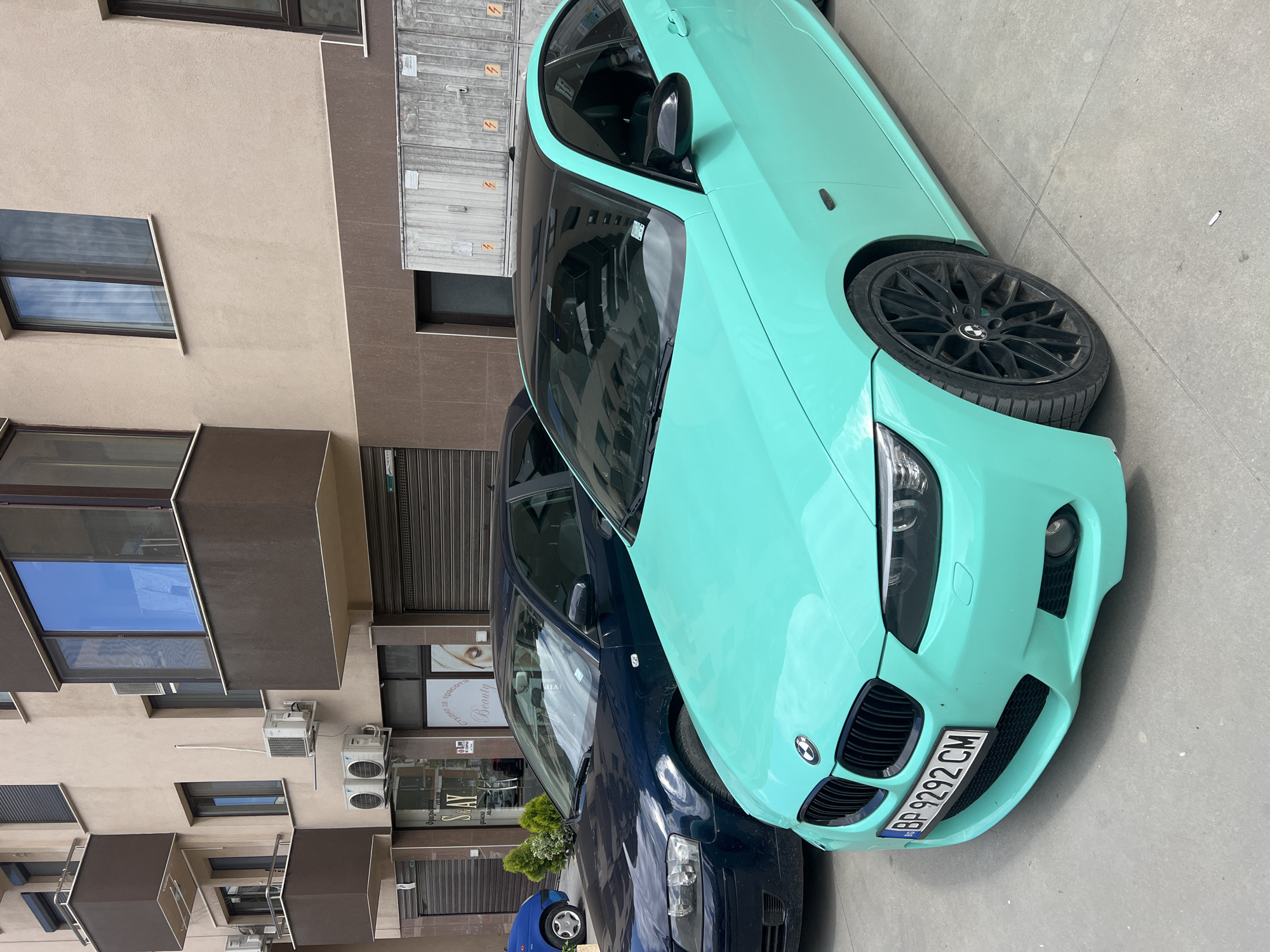 The image depicts a vibrant daytime urban street scene in Plovdiv, Bulgaria. The main focus is a striking mint green (or turquoise) BMW 3-series coupe, likely an E92 generation, parked on the paved ground. This car features distinctive black multi-spoke alloy wheels, a black front grille, and a contrasting black roof, creating a two-tone effect. Its Bulgarian license plate, "BP 9292 CM," is prominently displayed on the front bumper.
Behind the mint green BMW, partially obscured, is a dark blue or black sedan, appearing to be another German vehicle, possibly an Audi or an older BMW model. Further in the background, a small blue car's wheel and part of its body are visible.
The cars are parked alongside a multi-story building that forms the backdrop. The building has a light beige facade, possibly stucco or painted concrete, interspersed with darker brown-grey sections. Numerous rectangular windows with dark frames are visible, some reflecting the bright sky. The ground floor features commercial storefronts, some with closed roll-down metal shutters. Several outdoor air conditioning units are mounted on the building's exterior. Visible text on a storefront sign includes parts of the word "Beauty" and another stylized word, possibly "SEAY" or "SOAY," though some text is mirrored due to reflections. The scene suggests a typical, sunny day in a city environment.