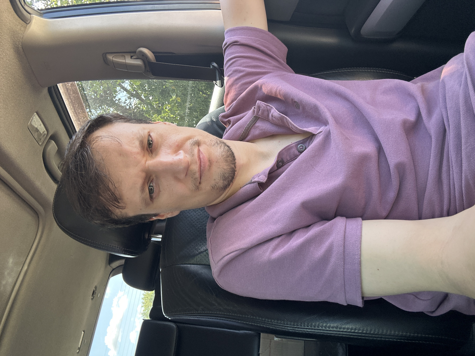 A young man with brown hair and a light complexion is seated in the driver's seat of a vehicle. He is wearing a purple polo shirt. His expression is a slight grimace, with his eyebrows furrowed. The interior of the car is visible, including the black leather seats, the sunroof, and a portion of the dashboard. Outside the car, through the windows, trees and a cloudy sky can be seen. The image appears to be a selfie, taken by the man himself. It is not possible to determine the specific location within Makiv, Ukraine, from the visual information provided.